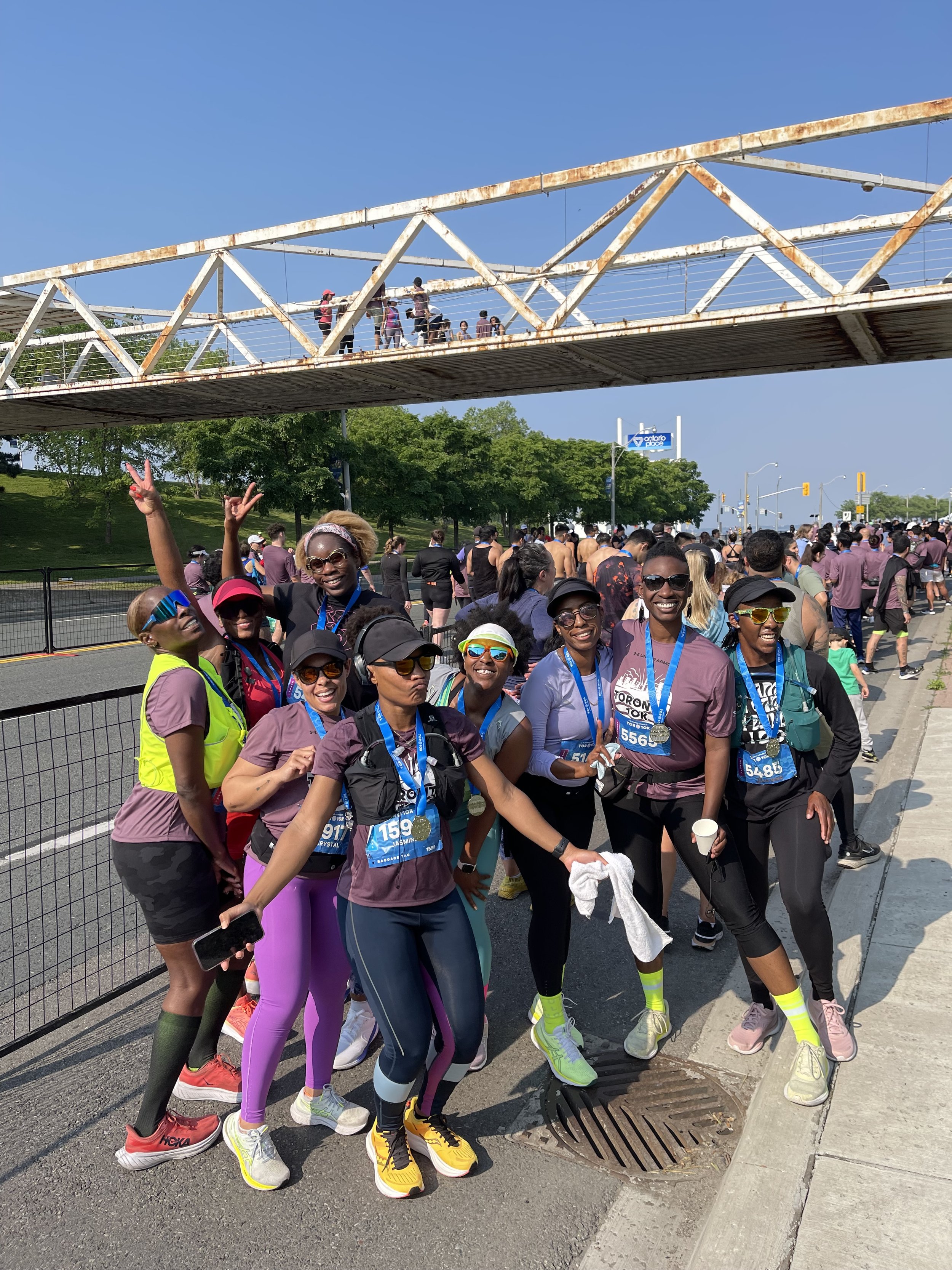 A group of Black women marathon runners celebrating after finishing a race, wearing medals and bright athletic gear, with others in the background and a bridge overhead.