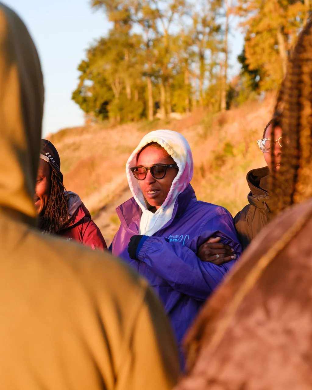Group of people standing outdoors during golden hour, with trees and a hillside in the background. The central person is wearing a purple jacket, white hoodie, and glasses, with eyes closed and hands crossed.