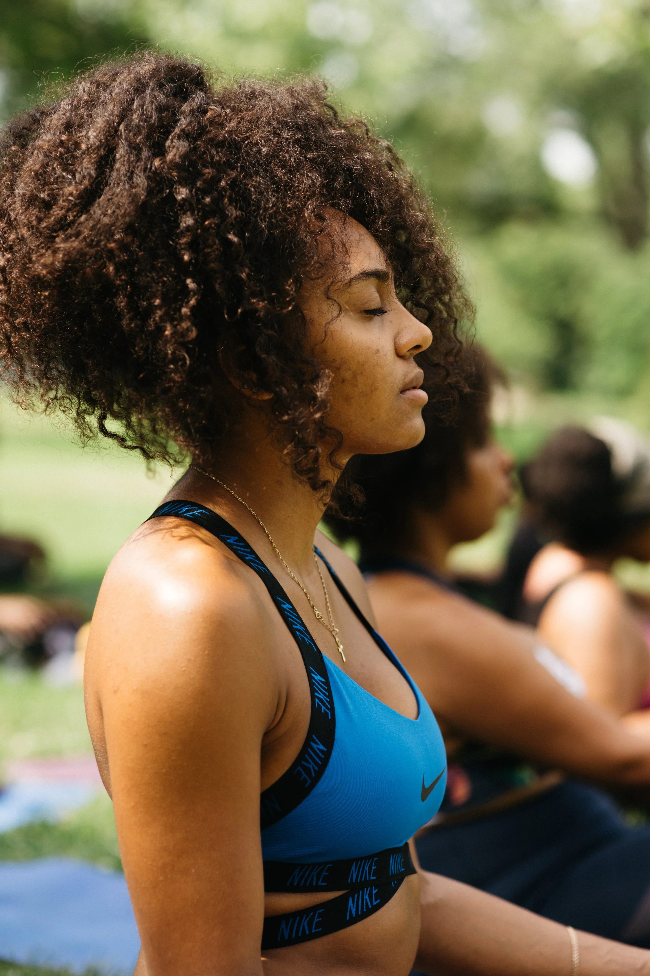 A young woman with curly hair practicing meditation outdoors, sitting cross-legged with her eyes closed, wearing a blue Nike sports bra and a gold cross necklace.