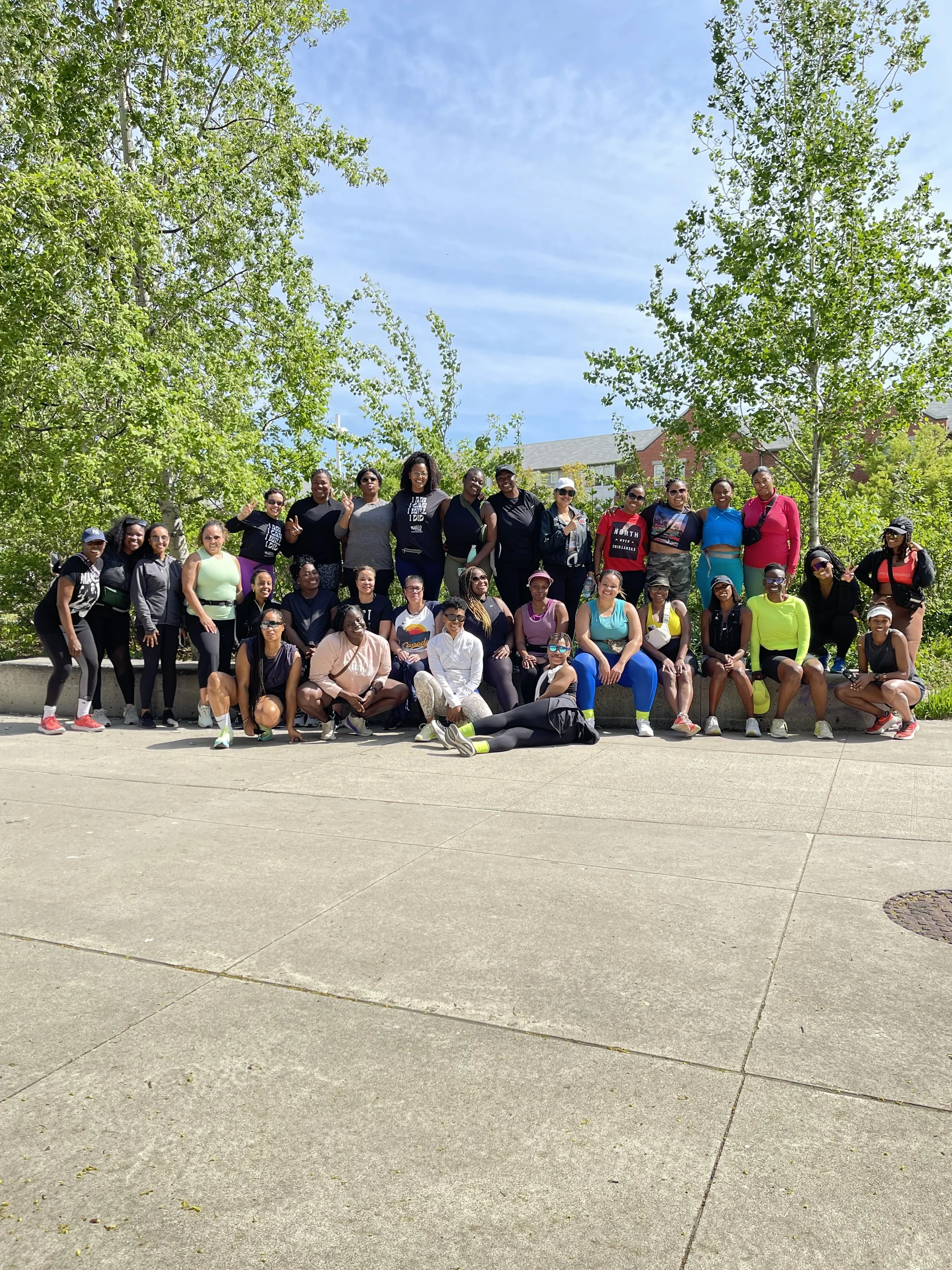 Group of diverse women posing outdoors on a sunny day, with green trees and a blue sky in the background.