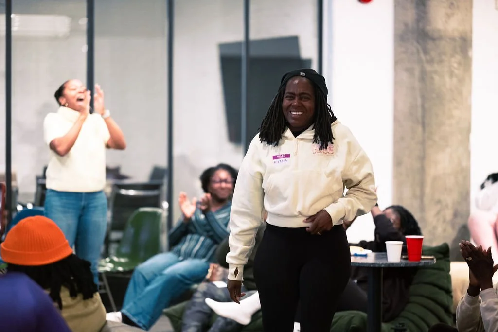 A group of women, one in the foreground smiling and wearing a beige jacket and a name tag, in a room with some chairs and tables, people clapping and laughing.