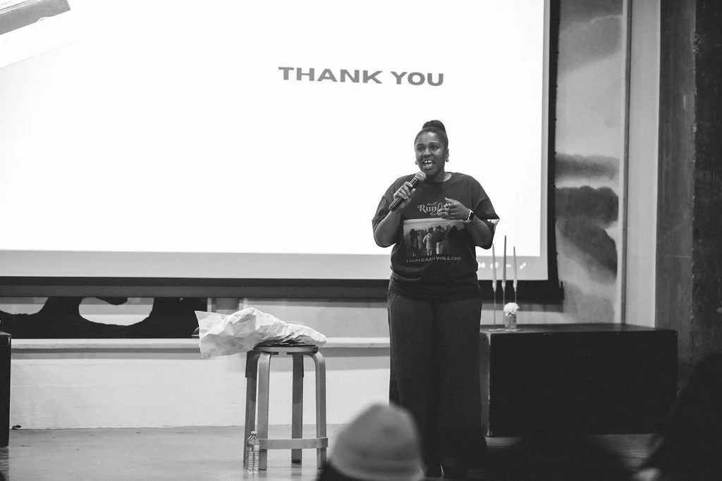 A woman standing on stage with a microphone in hand, smiling and speaking to an audience, in front of a screen that says 'Thank You.'