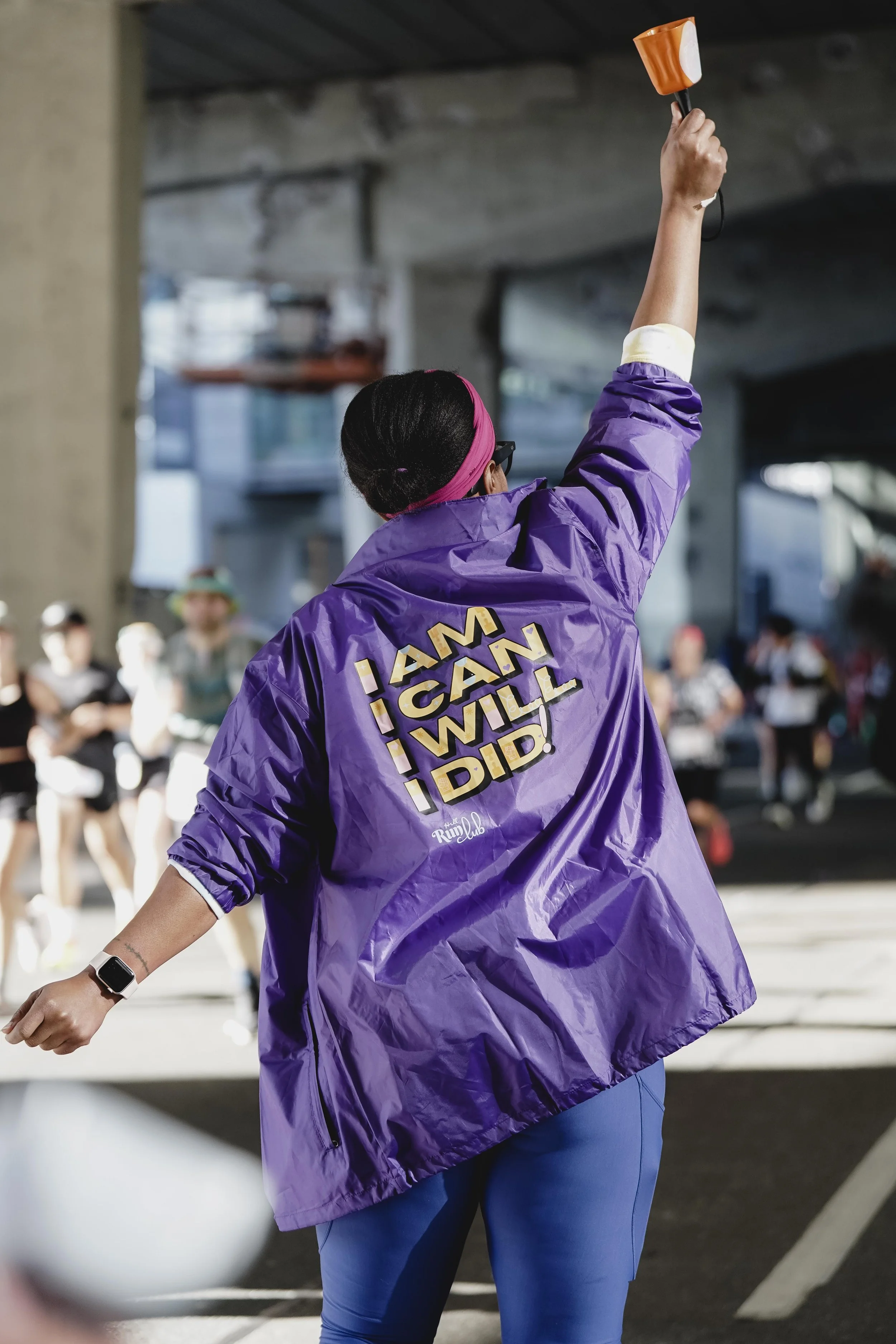 A woman wearing a purple jacket with the words 'I AM, I CAN, I WILL, I DID' on the back, participating in a fitness event or race under a bridge.