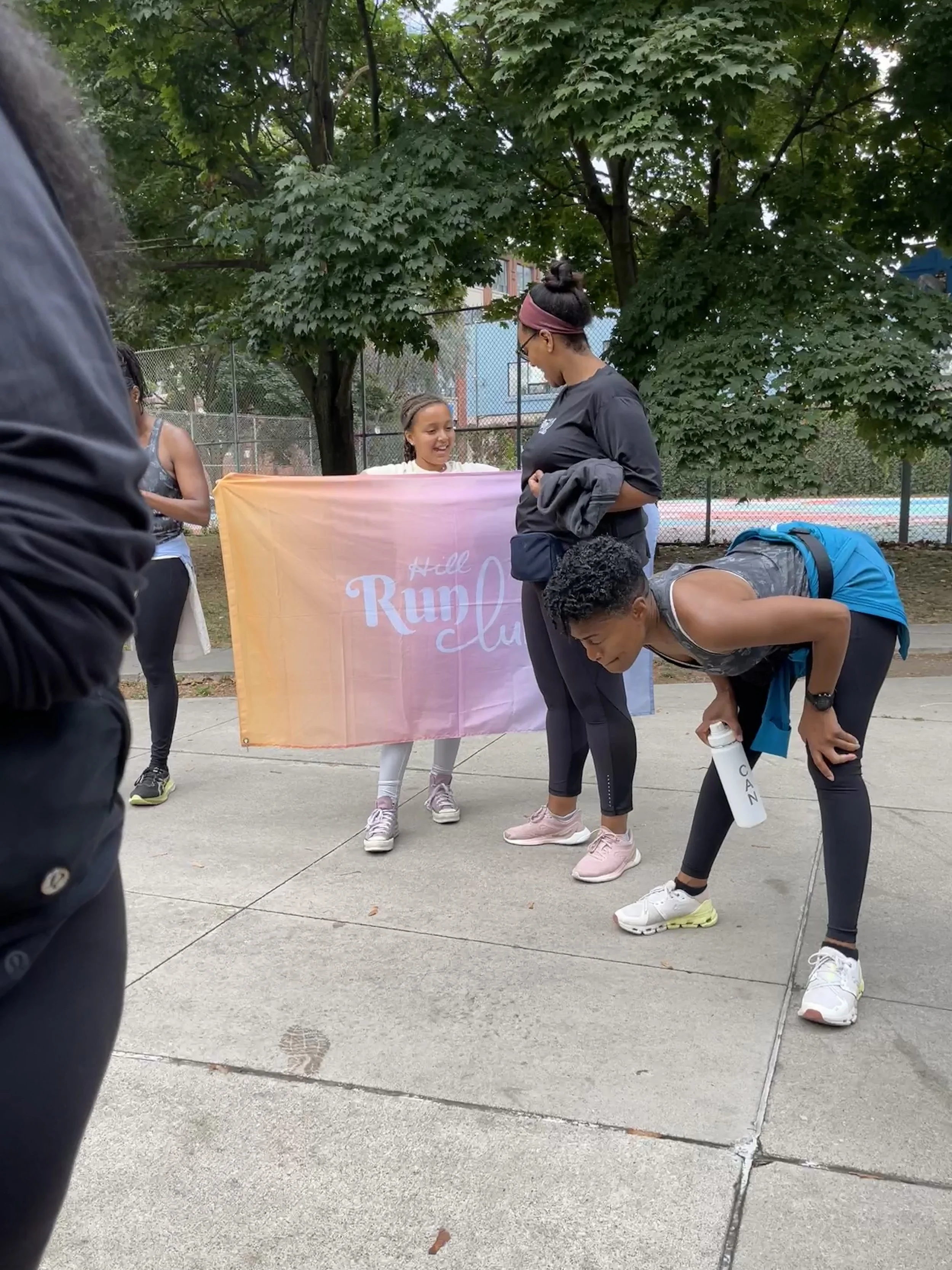 Group of runners, mostly women, preparing at a park for a race, with one woman drinking water, a girl smiling behind a banner that reads 'Hill Run Club,' and others getting ready to start.