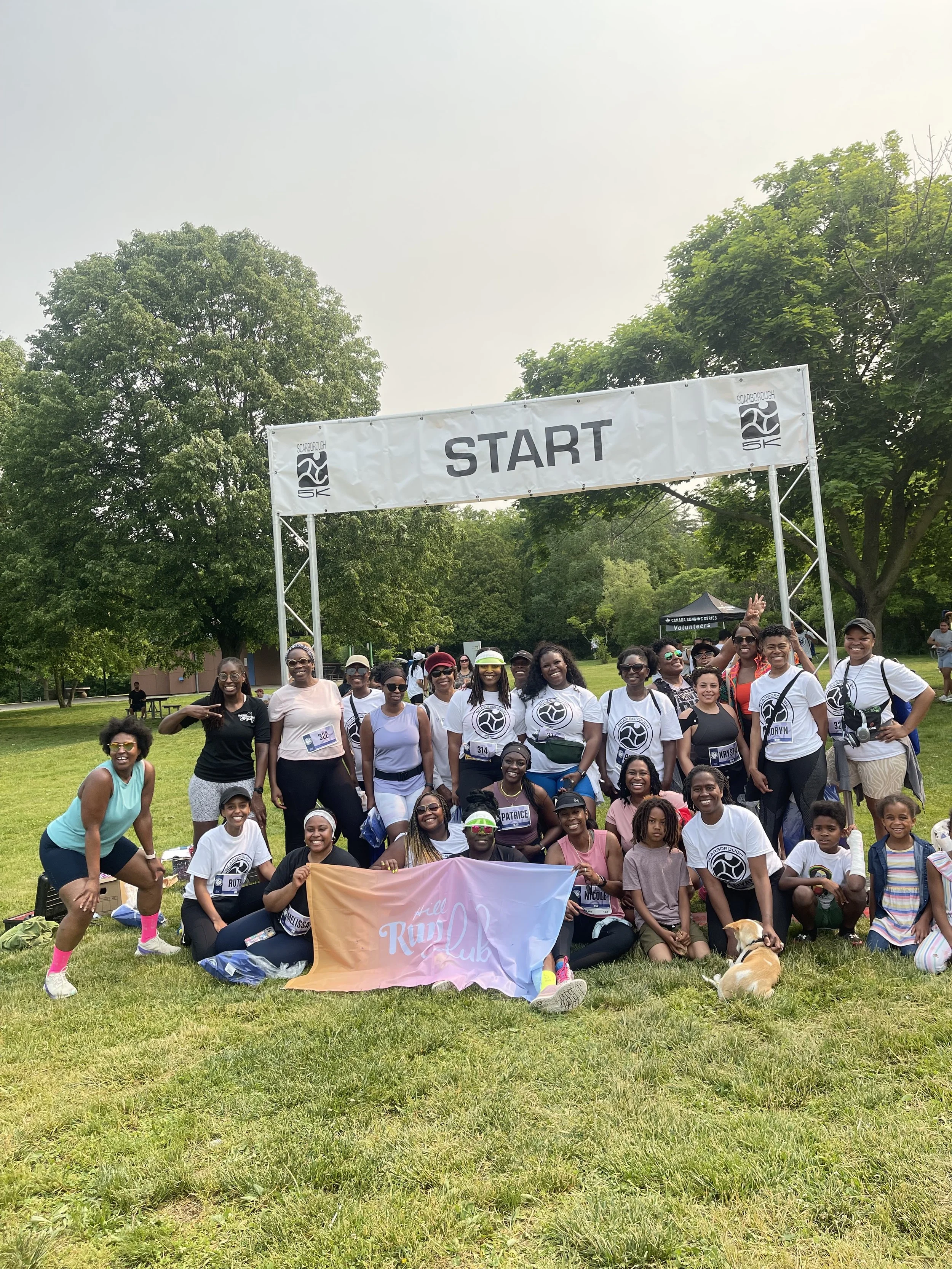 Group of people gathered outdoors near a start line banner for a running event, with some holding a pink banner that reads 'Run Club,' and a dog lying on the grass in front.