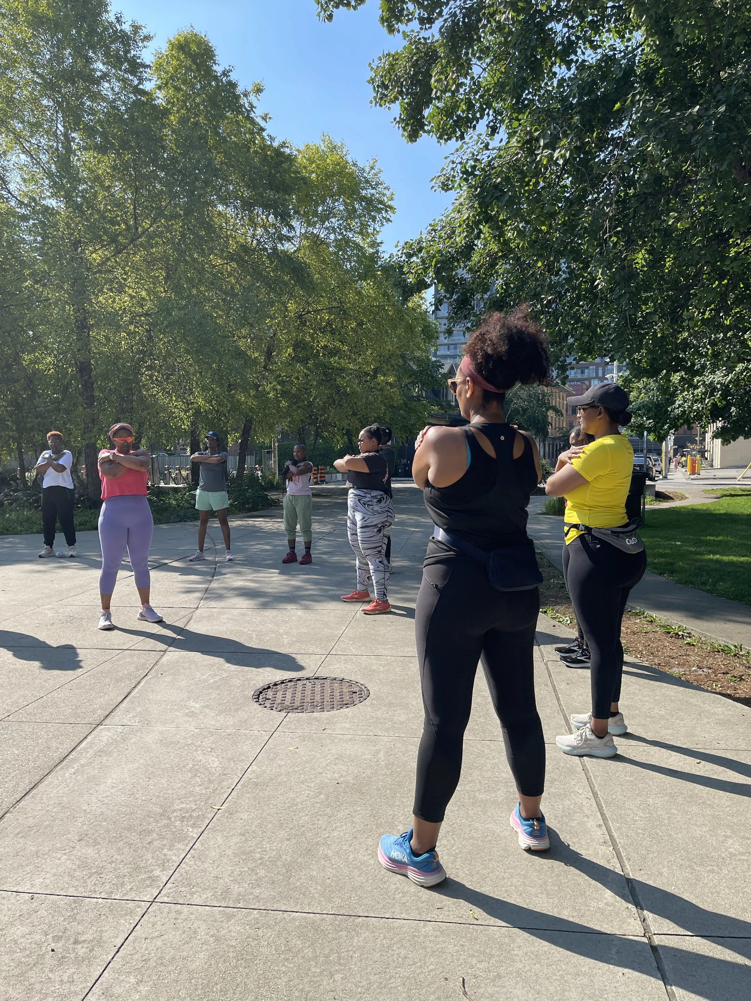 A group of women participating in an outdoor exercise class in a park, standing on a concrete sidewalk with trees and buildings in the background.