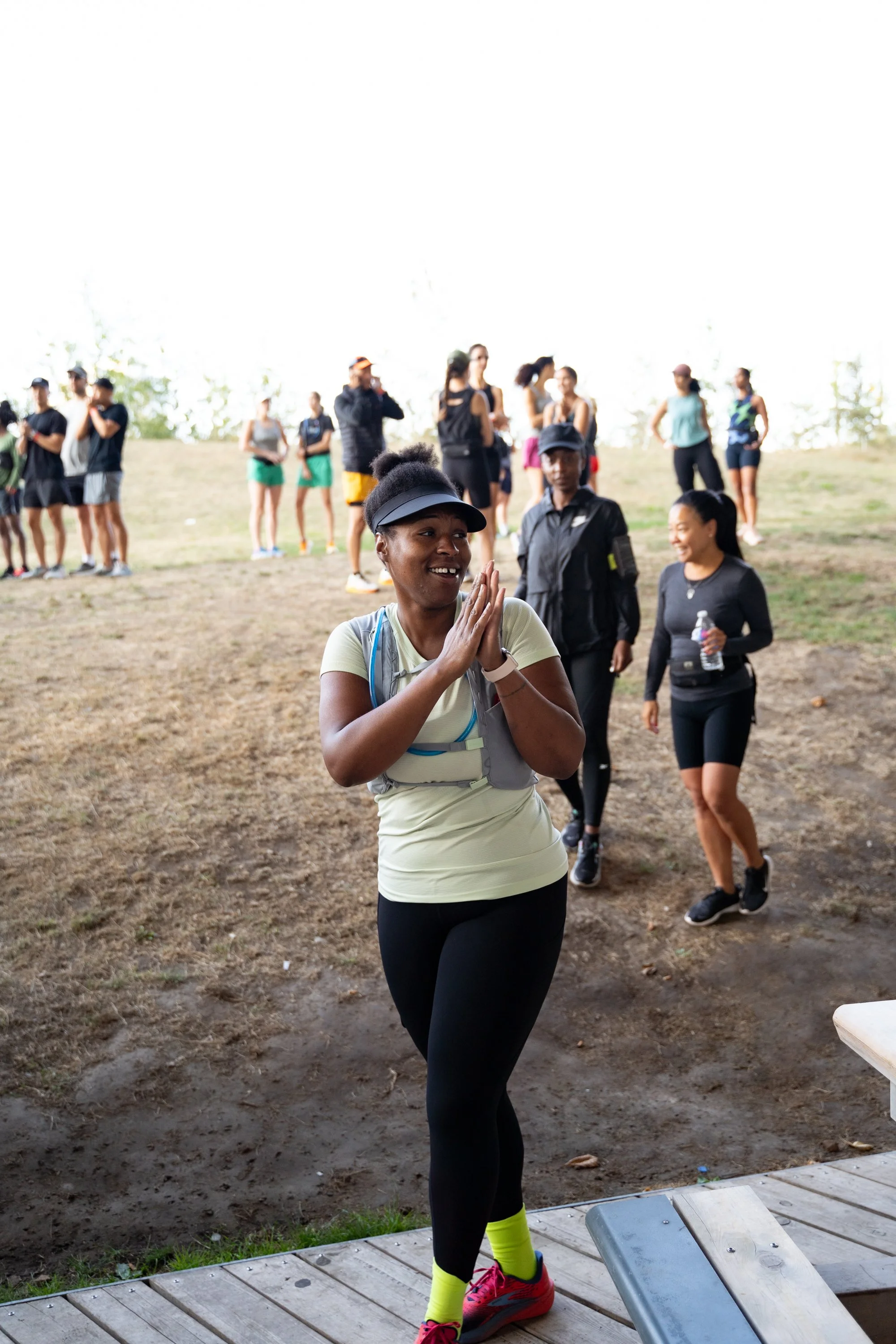 A woman in athletic clothing and a visor smiles with her hands pressed together as she approaches the camera. Other runners and one security guard are visible in the background at an outdoor event.