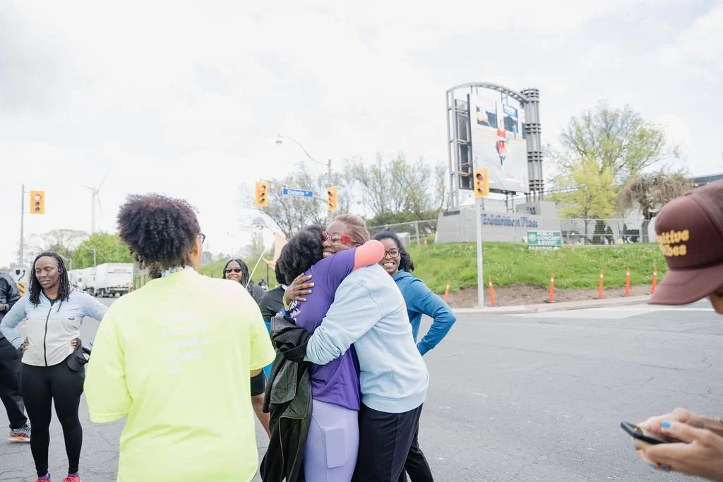 Group of people hugging and smiling outdoors in a parking lot, with traffic lights and a billboard in the background.