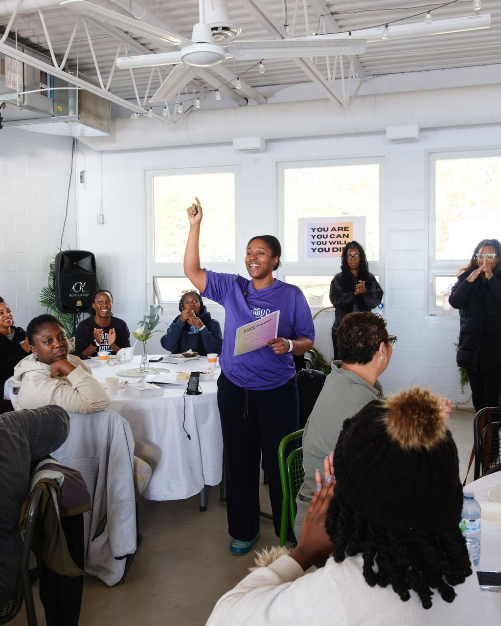 A woman standing and speaking to a group during a gathering or workshop, with others seated around a table, some smiling and clapping in a bright, modern room.