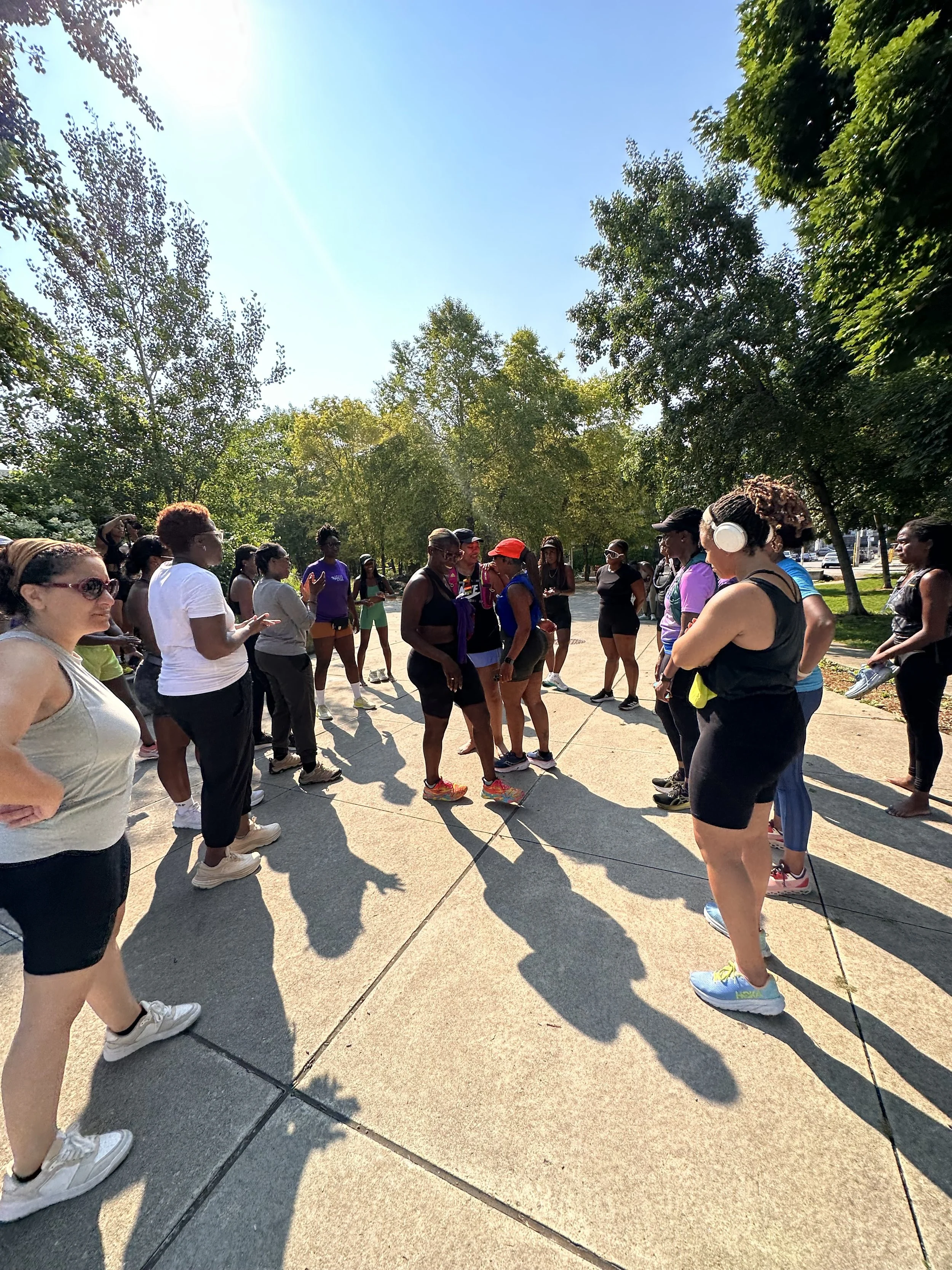 Group of women gathered outdoors in a park-like area on a sunny day, some wearing headphones, engaging in conversation or listening, with trees and a blue sky in the background.