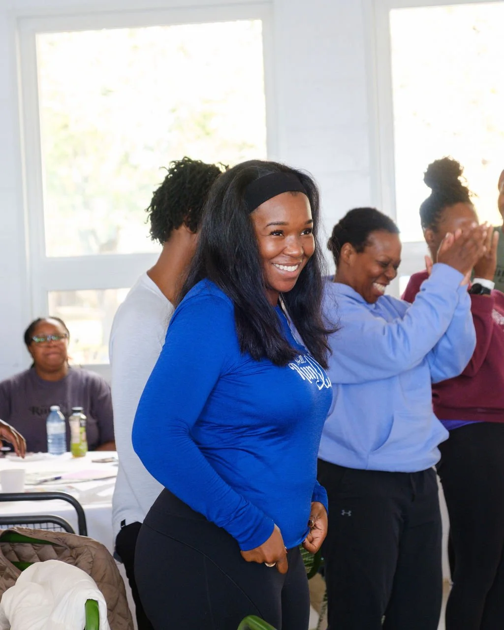 Group of women smiling and laughing, standing indoors near windows, with one woman in blue in the foreground.
