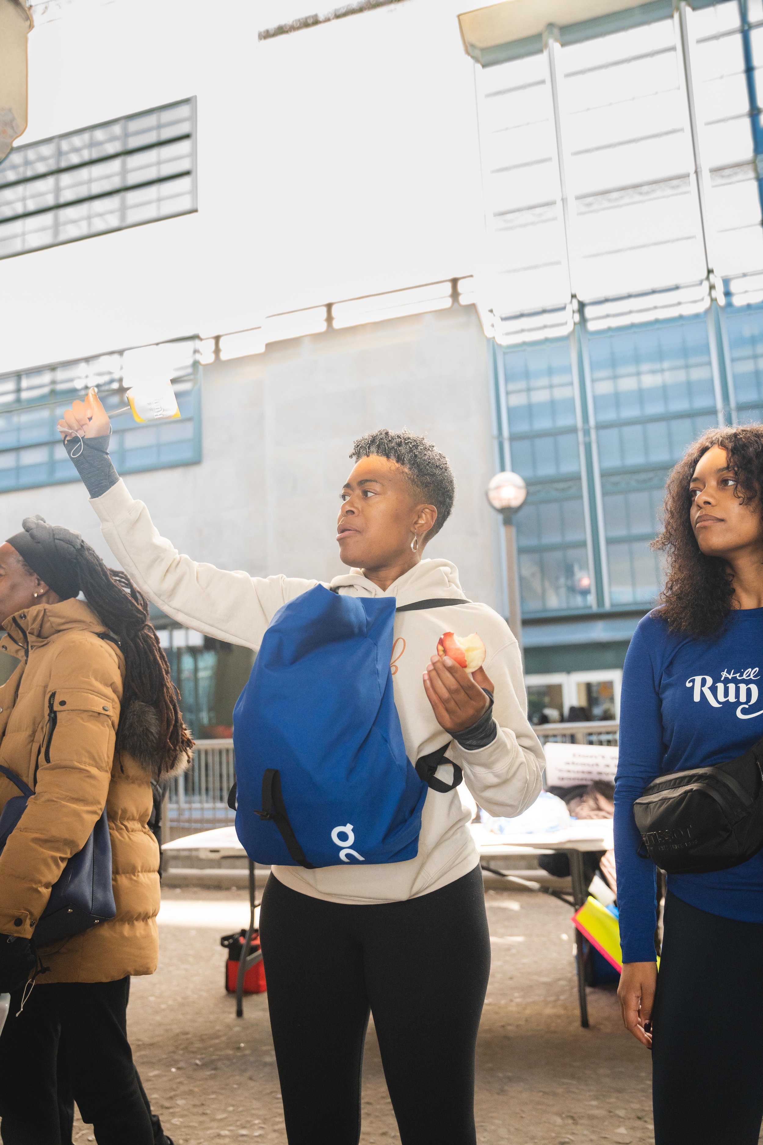 Group of women standing outside in an urban area, some holding apples, one with a blue backpack, some wearing hoodies and jackets, at a table with papers in the background.