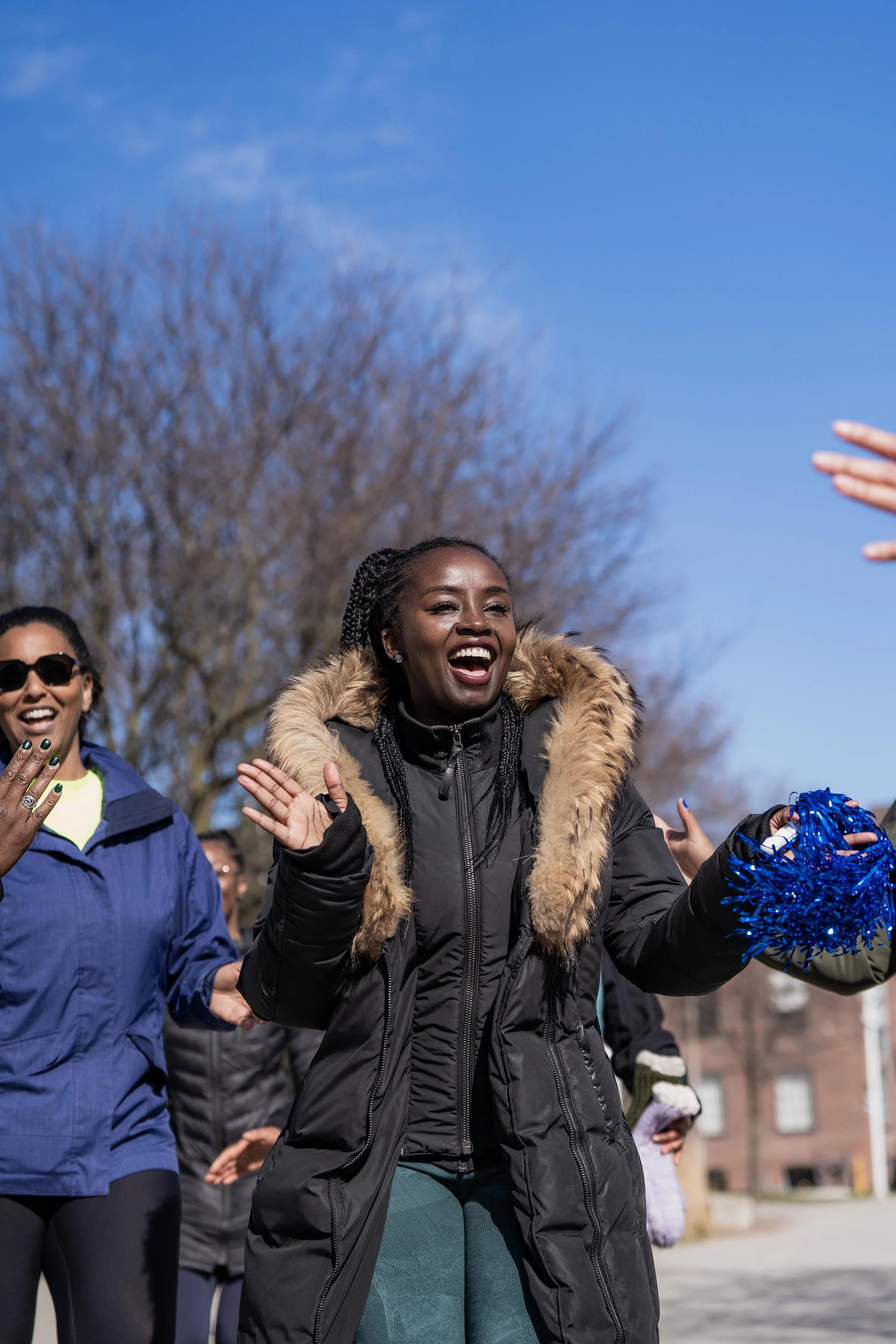 Women participating in an outdoor public celebration or event, some clapping, under a clear blue sky with leafless trees in the background.