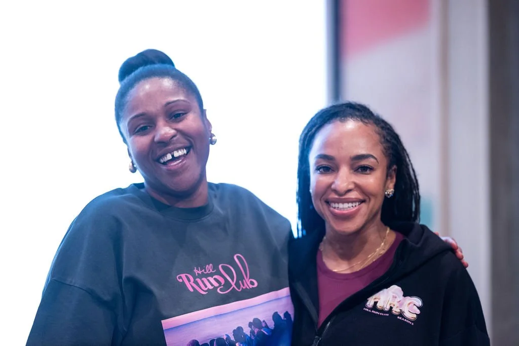 Two smiling women posing indoors, one wearing a black shirt with pink text 'Hill Run Club', and the other wearing a black zip-up sweater with a butterfly logo.