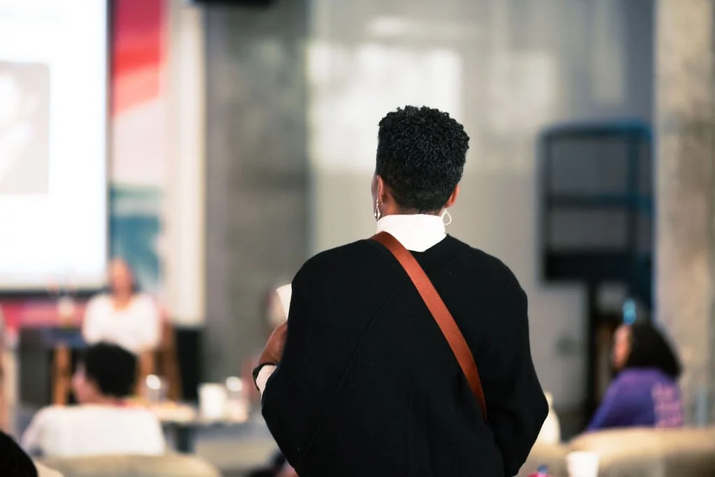 Person with short curly hair, wearing a black sweater and white collared shirt, attending a presentation or lecture in a conference room.