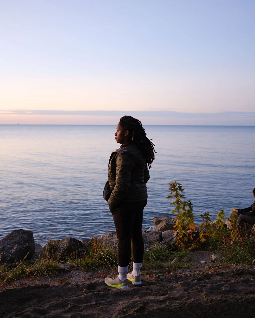 A woman with dreadlocks stands on a rocky shoreline, looking at the calm water and sunset sky.