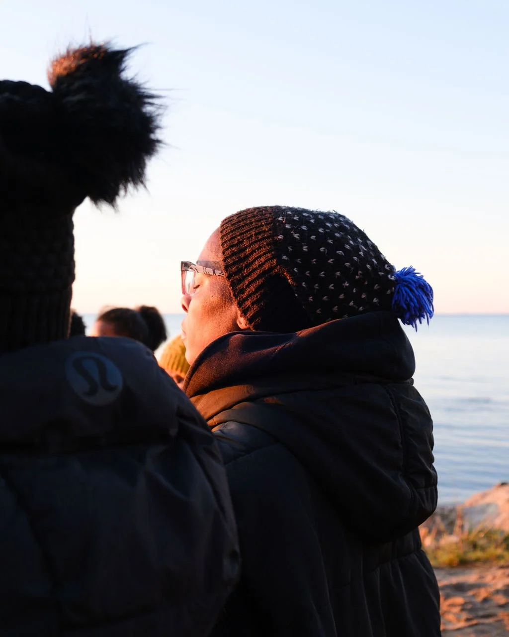 Person wearing glasses and a knitted hat with a pom-pom, standing near water during sunset, with other people in the background.