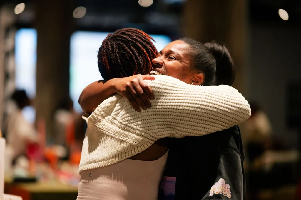 Two women are hugging and smiling in a warm embrace in an indoor setting.