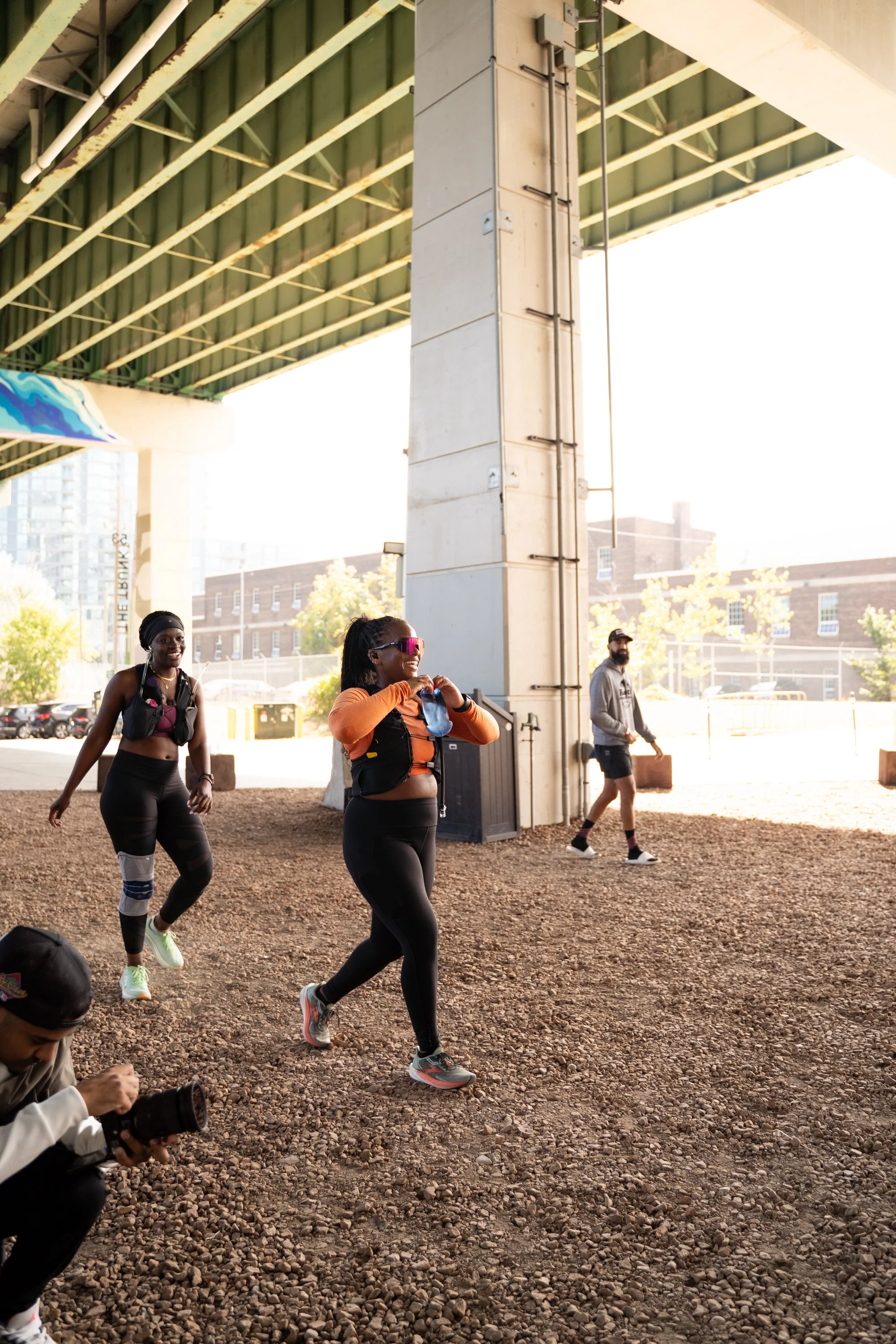 Three women dressed in athletic gear under a bridge, smiling and walking, while a man takes a photo near them.