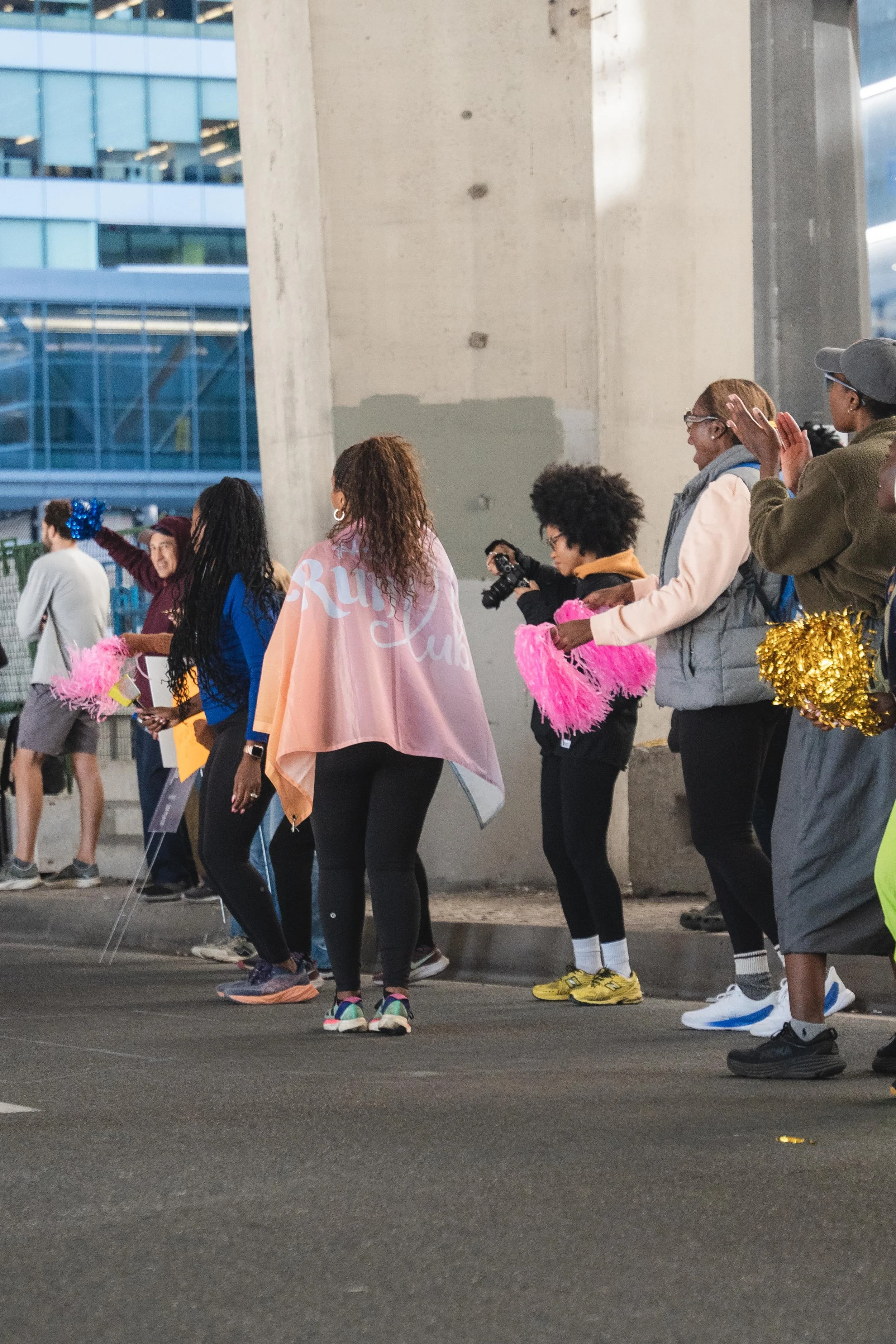 A group of women and a man standing on the street, holding pink and gold pom-poms, with some clapping and others talking. They are wearing casual clothing, and some are dressed warmly. In the background, there is a large concrete structure and modern