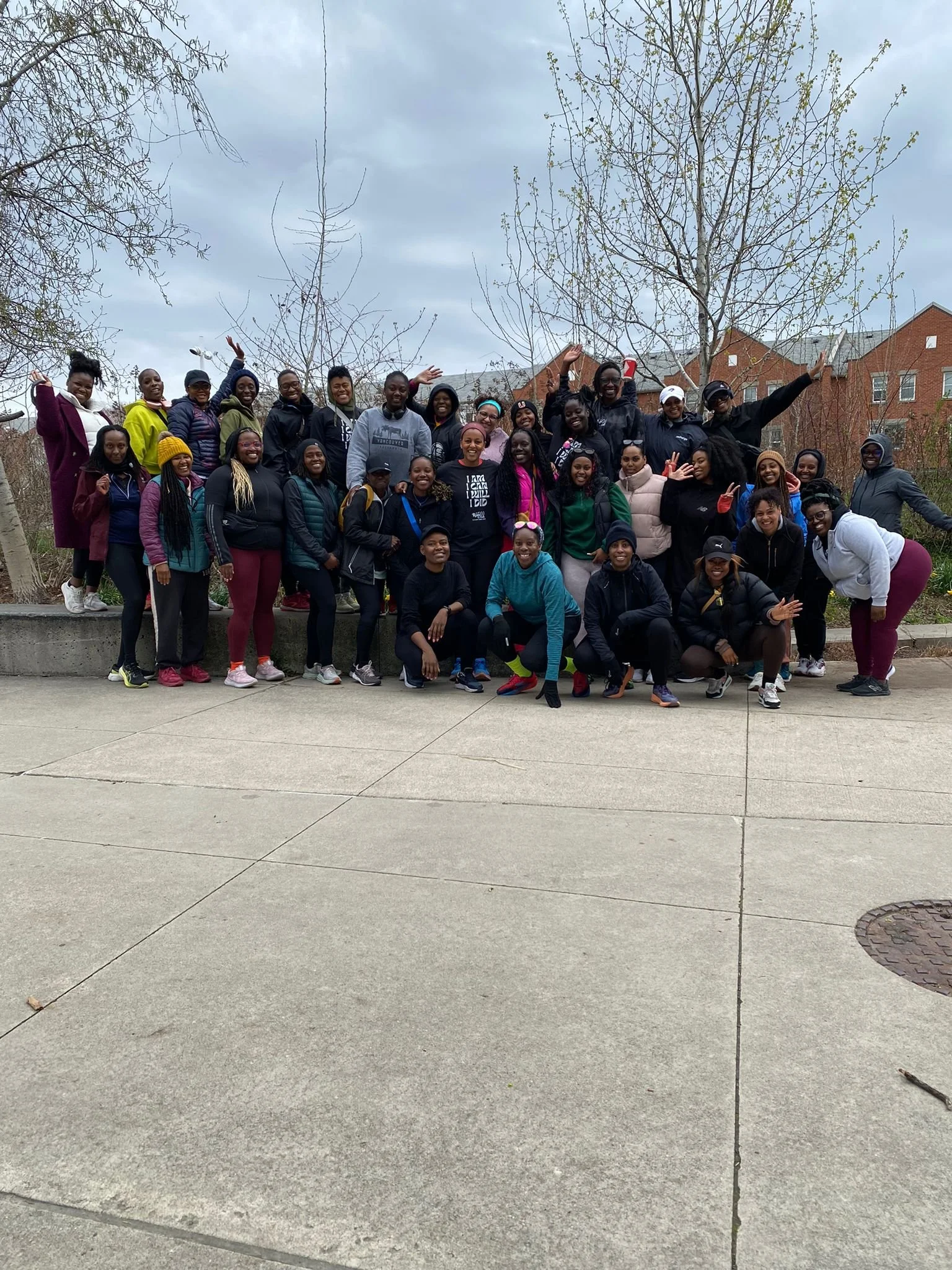 Group of people, diverse in age and ethnicity, posing outdoors on a cloudy day. They are standing and squatting on a sidewalk with leafless trees and houses in the background, smiling and waving.