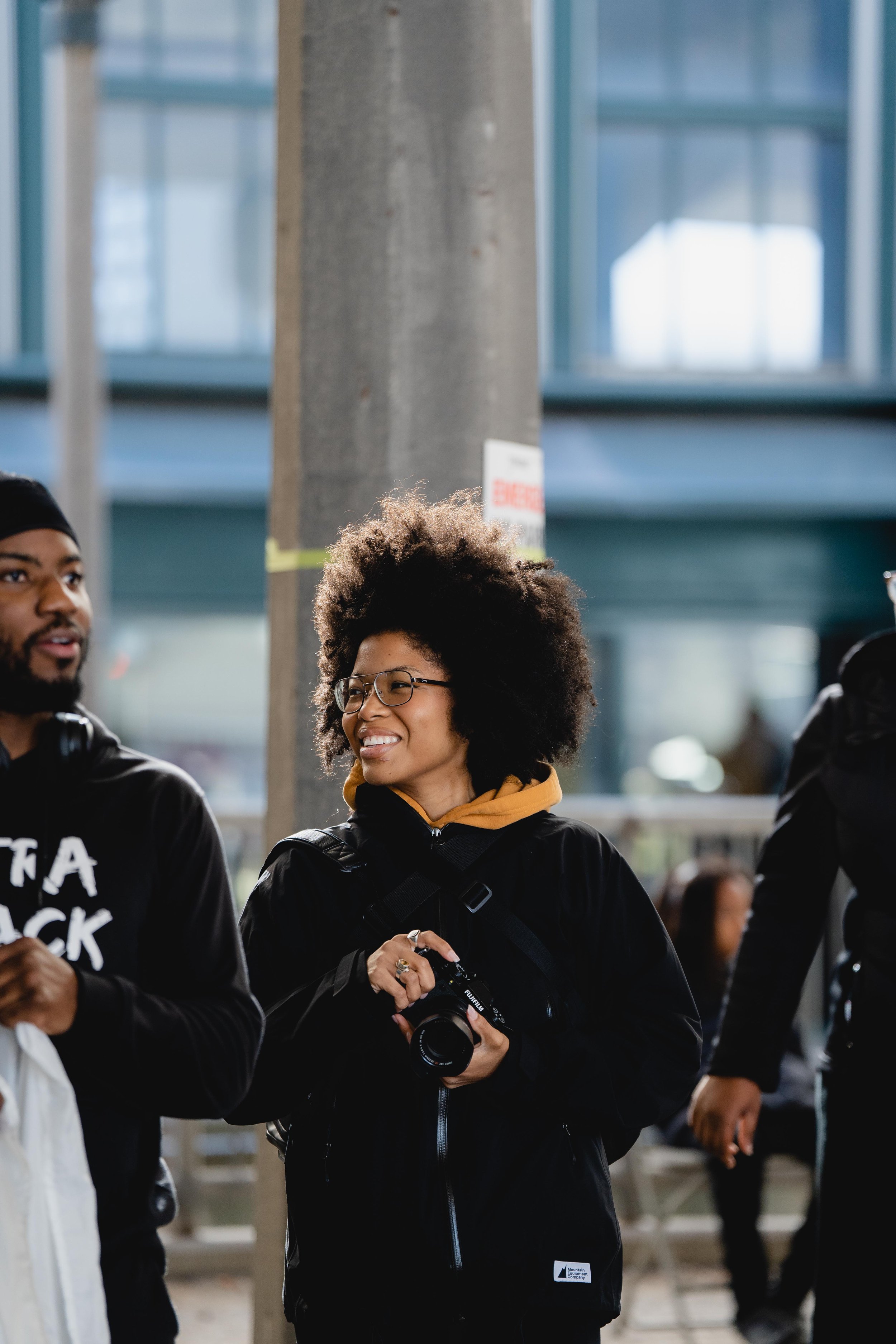 A woman with curly hair and glasses smiling, holding a camera, in a crowd with a man to her left and another person to her right in an industrial area with large windows.