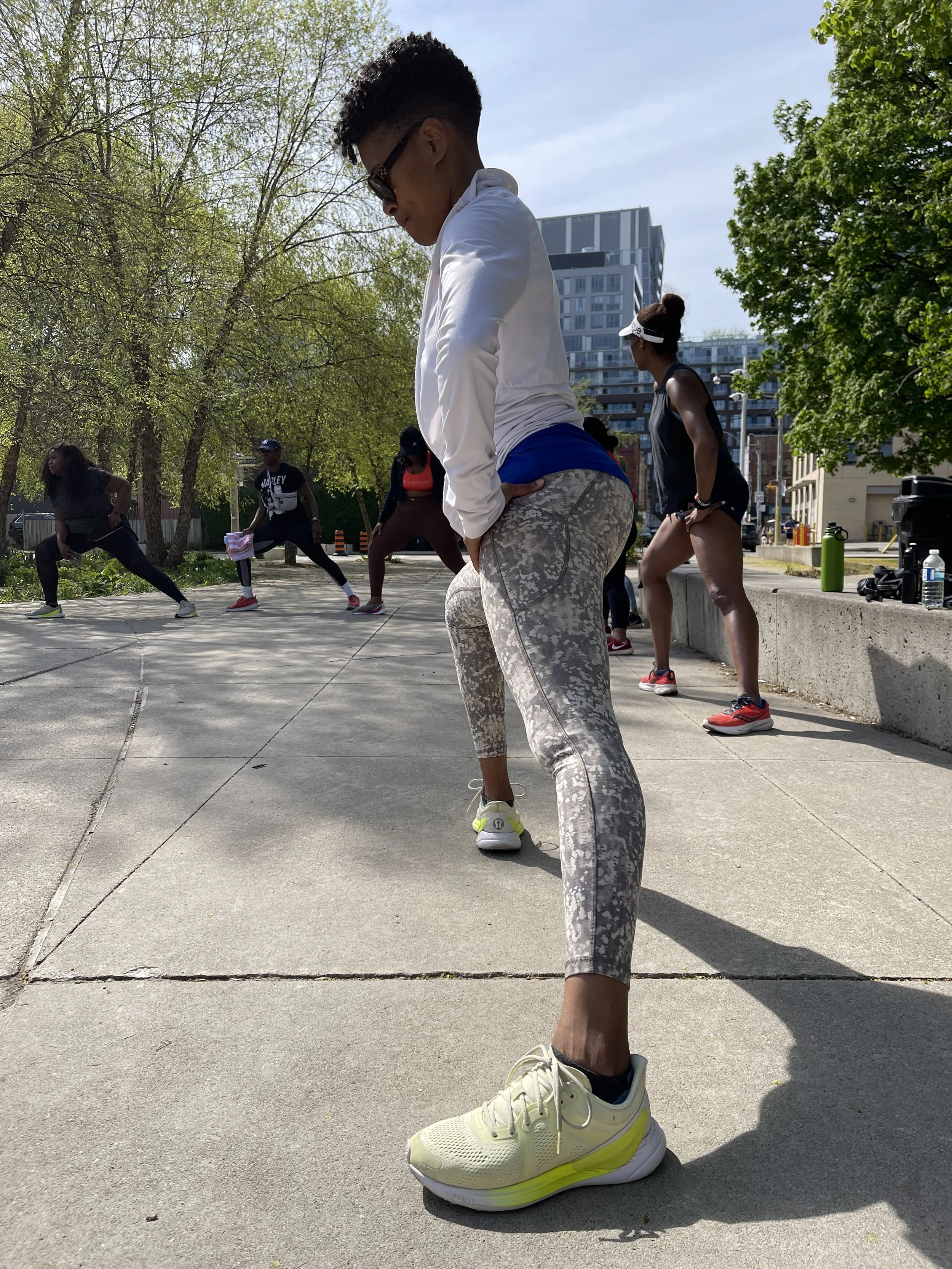 Group of women participating in outdoor fitness class, stretching on sidewalk in park with trees and city buildings in background.