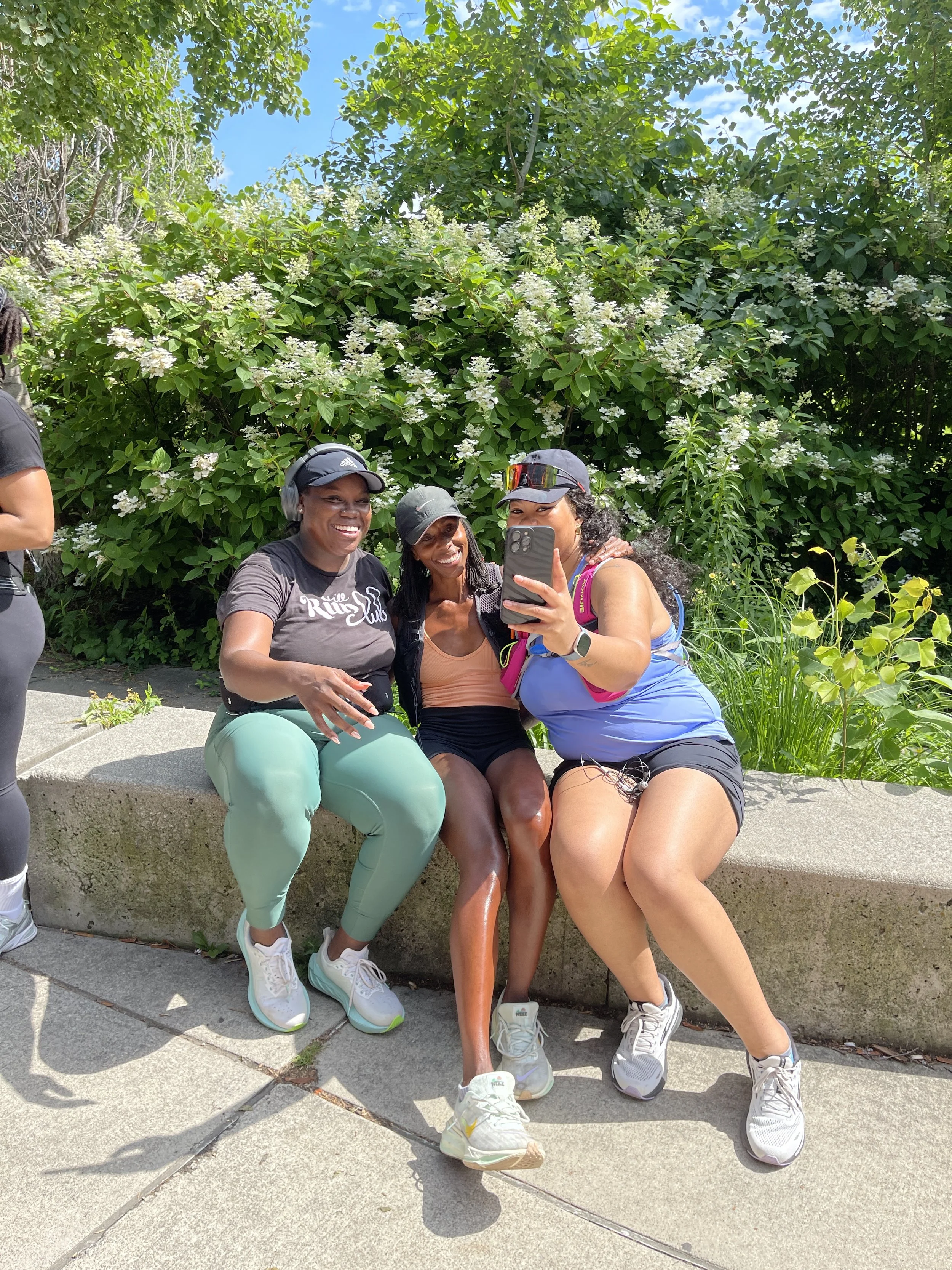 Three women sitting on a concrete ledge taking a selfie with a phone, surrounded by green foliage and blooming bushes.