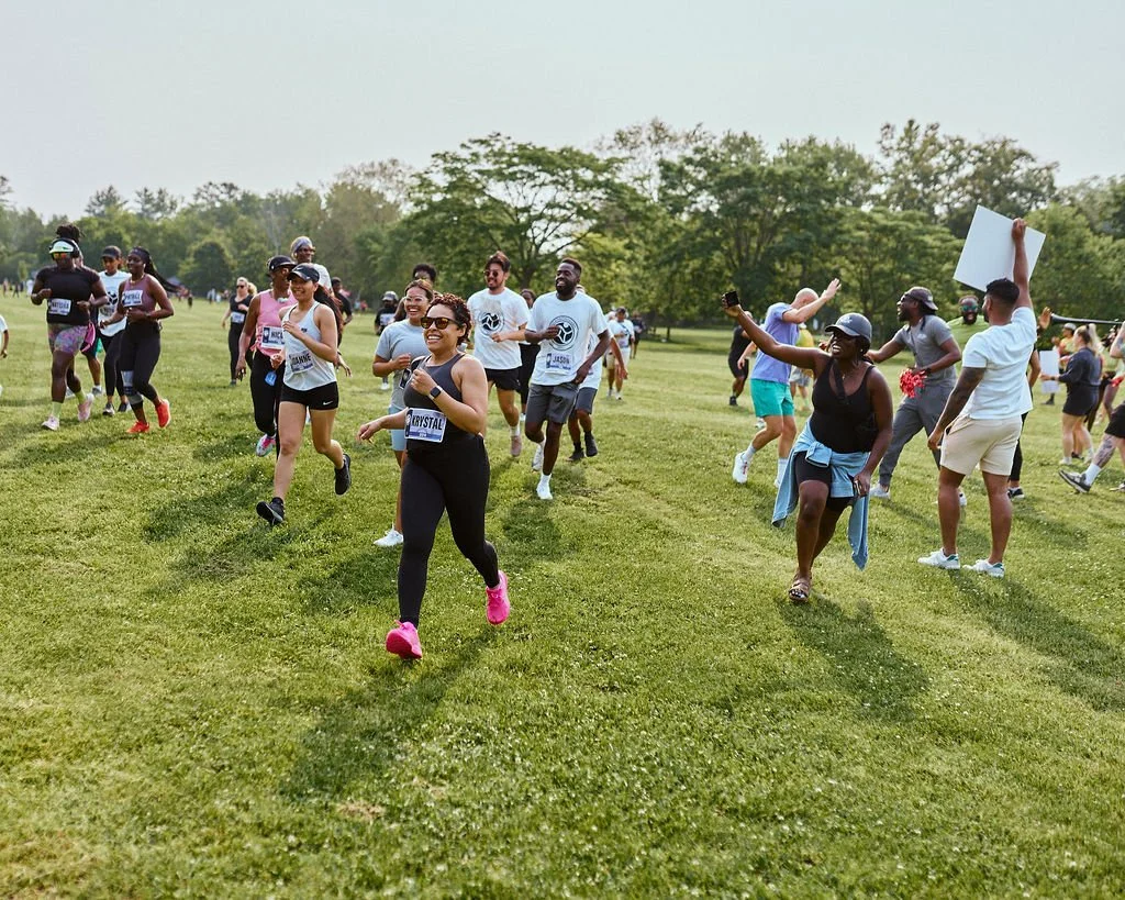 A group of people running and participating in a fun run or race on a grassy field, with some smiling and others cheering, during daytime with trees in the background.