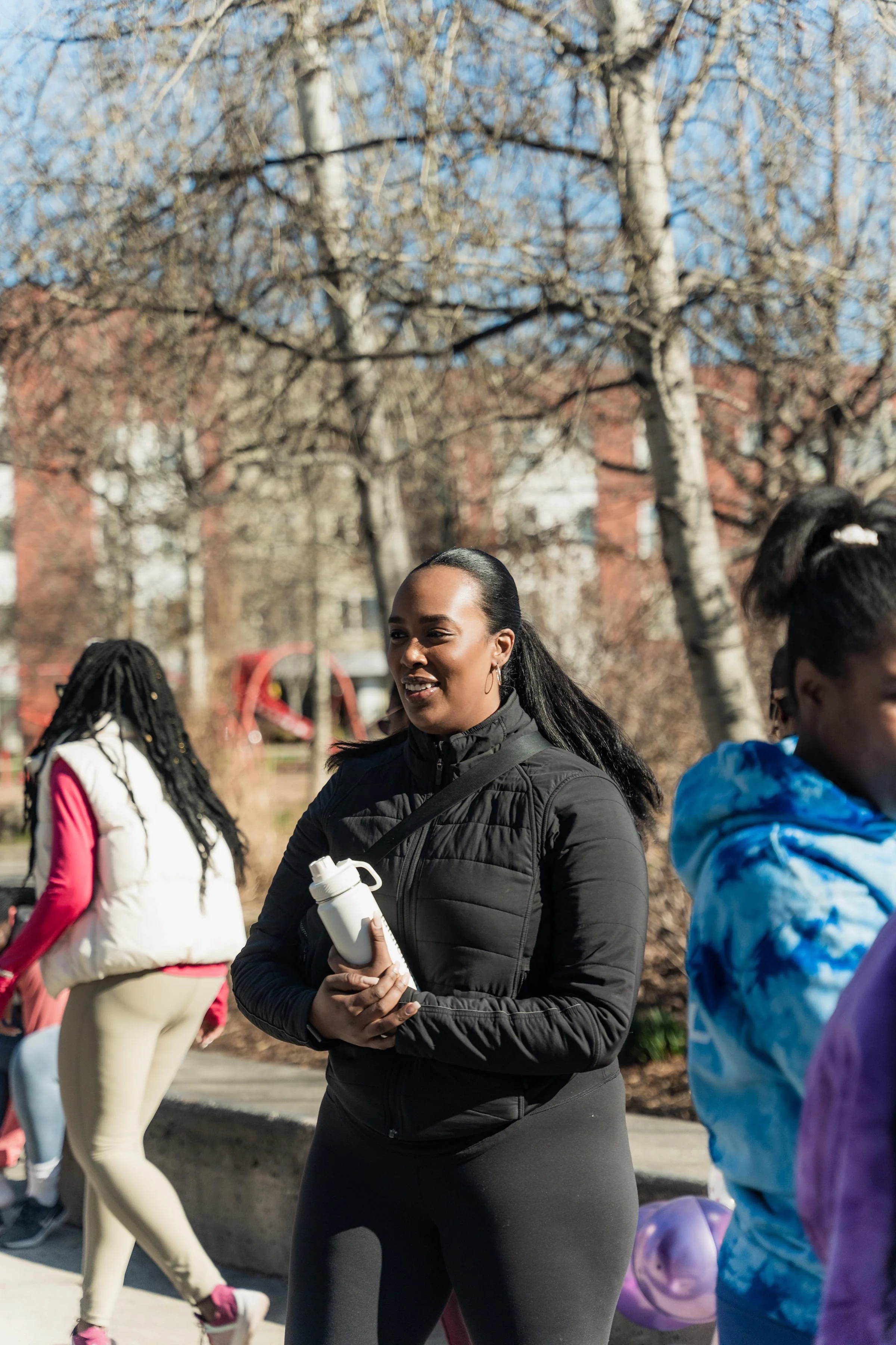 A woman in black athletic clothing holding a white water bottle, standing outdoors with other women in colorful jackets, trees, and buildings in the background.