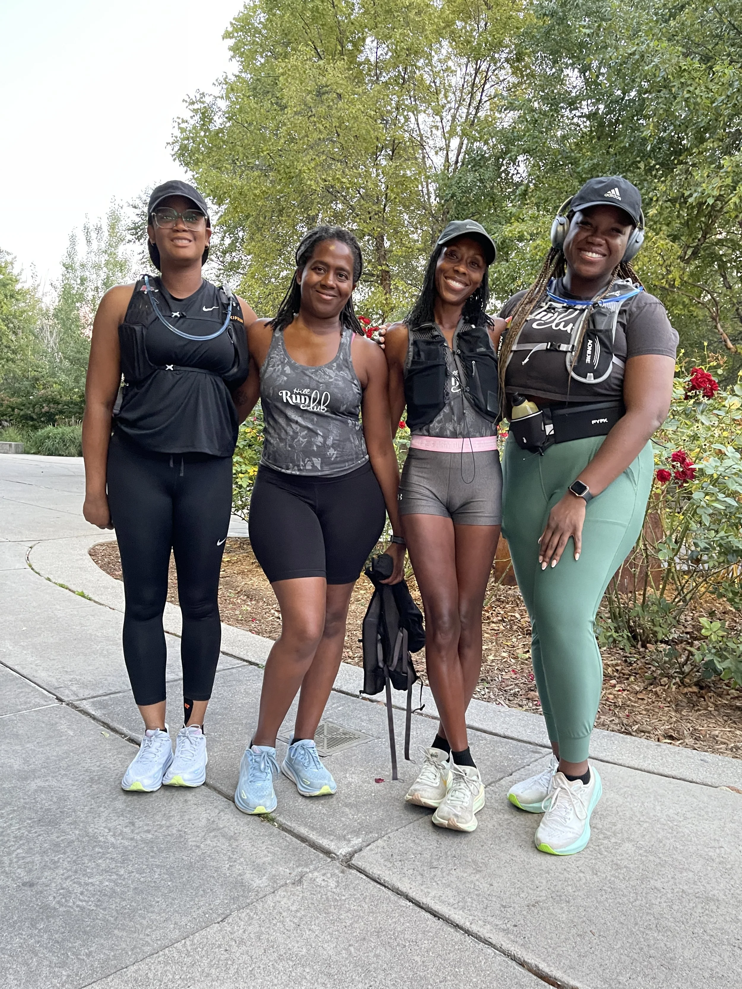 Four women in athletic gear standing outdoors on a sidewalk, smiling, with trees and bushes in the background.