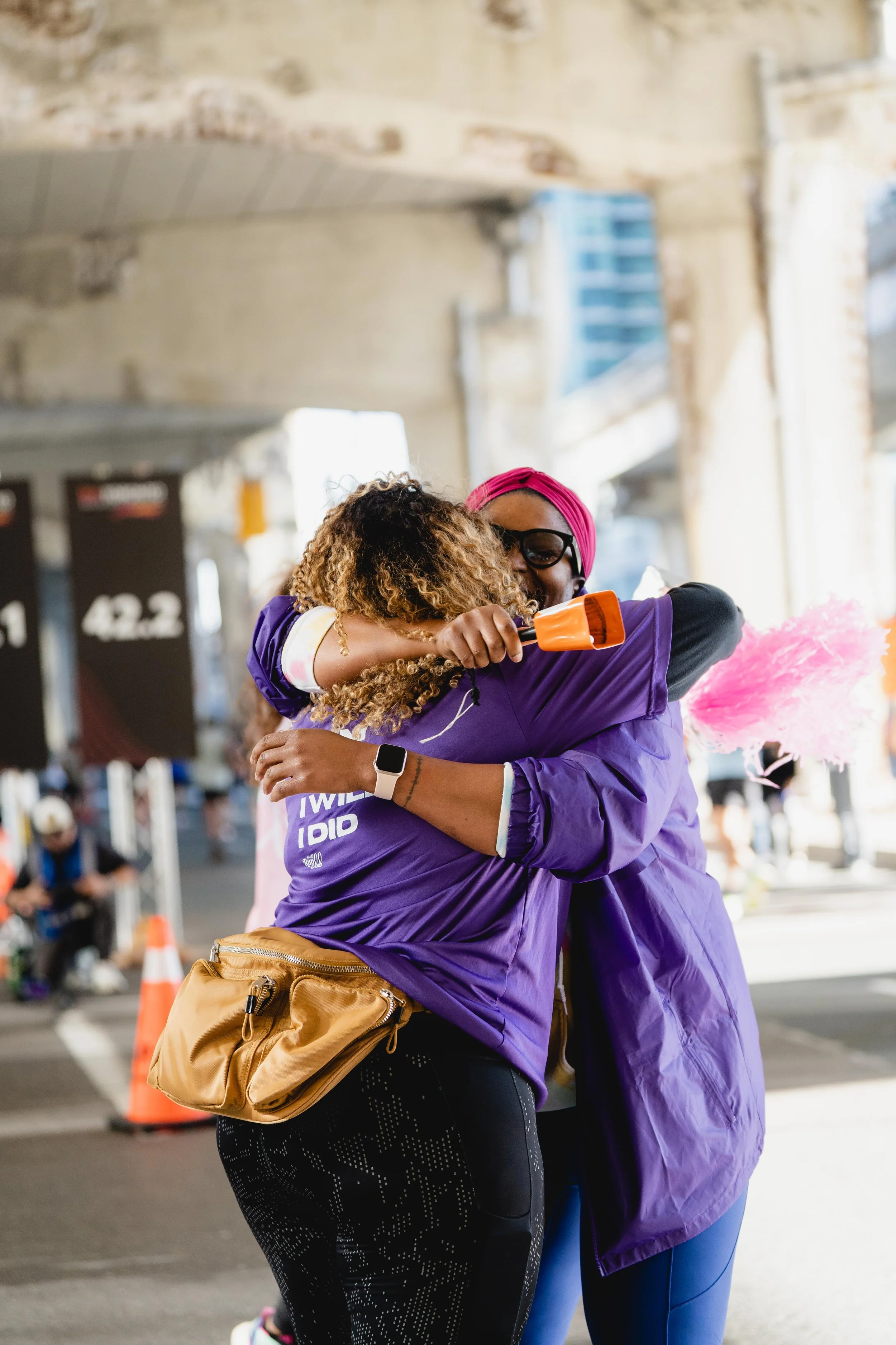 Two women hugging, one holding a pink smoke flare, under a bridge or overpass, at a race or event.