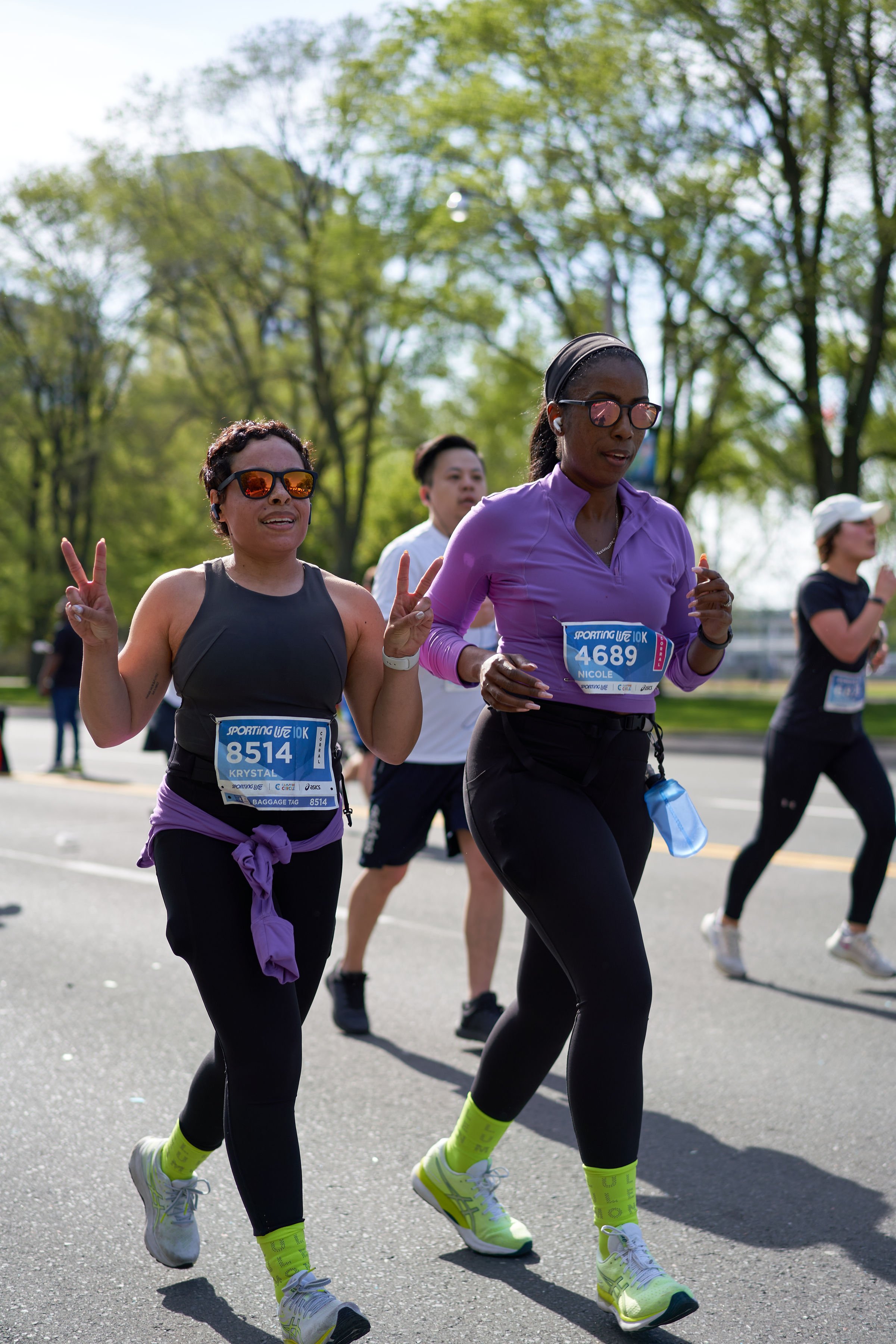 Two women running in a marathon, one making a peace sign, both wearing sunglasses and athletic gear, with other runners and green trees in the background.
