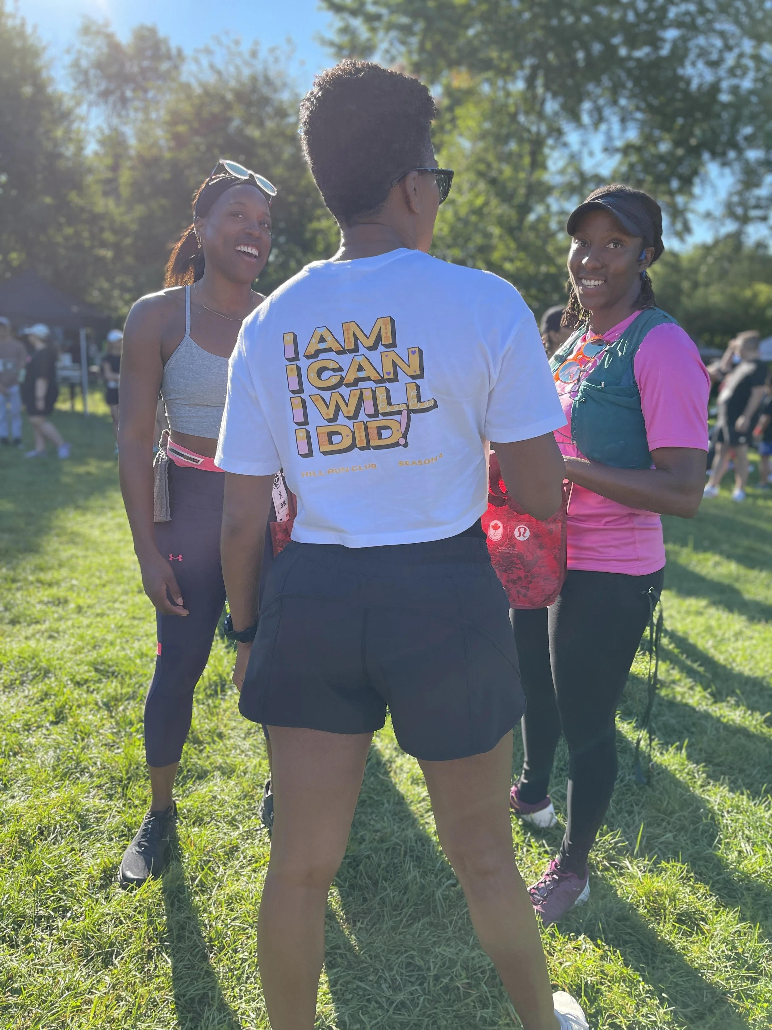 Three women are outdoors in a grassy area, engaged in conversation during a sunny day with trees in the background. The woman in the center has her back to the camera, wearing a white T-shirt with the words 'I AM, I CAN, I WILL, I DID!' printed on it