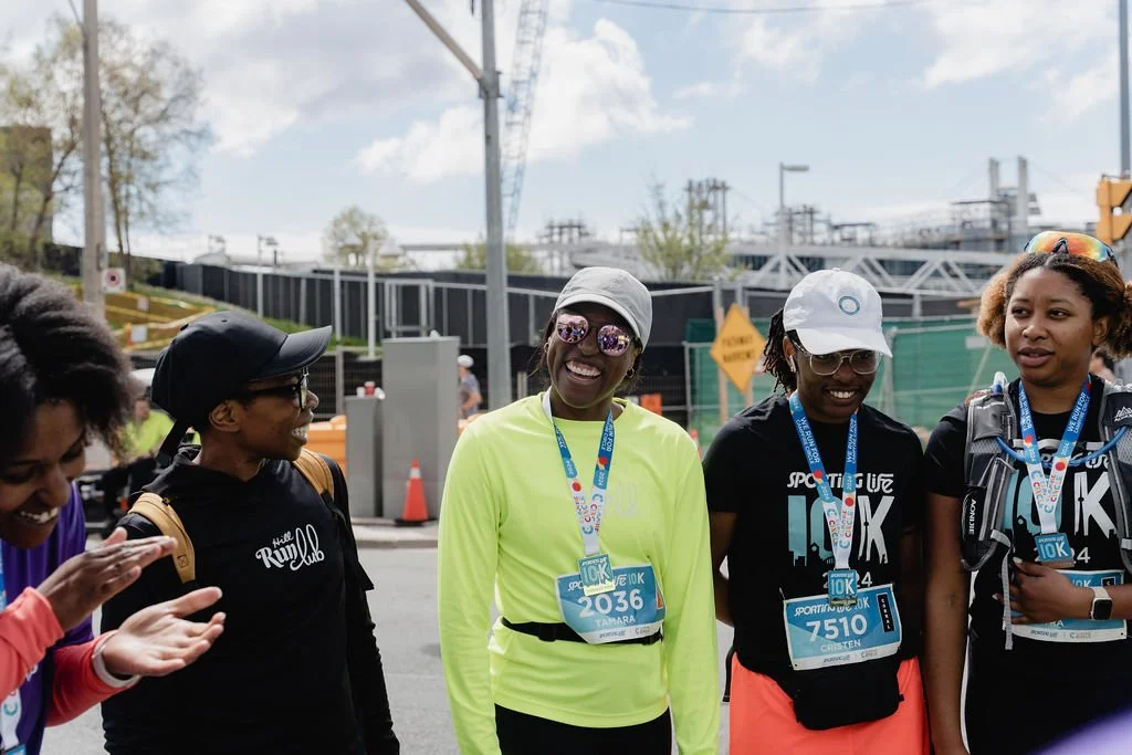 Group of women in running gear with race bibs, standing outdoors on a street after a 10K race event.