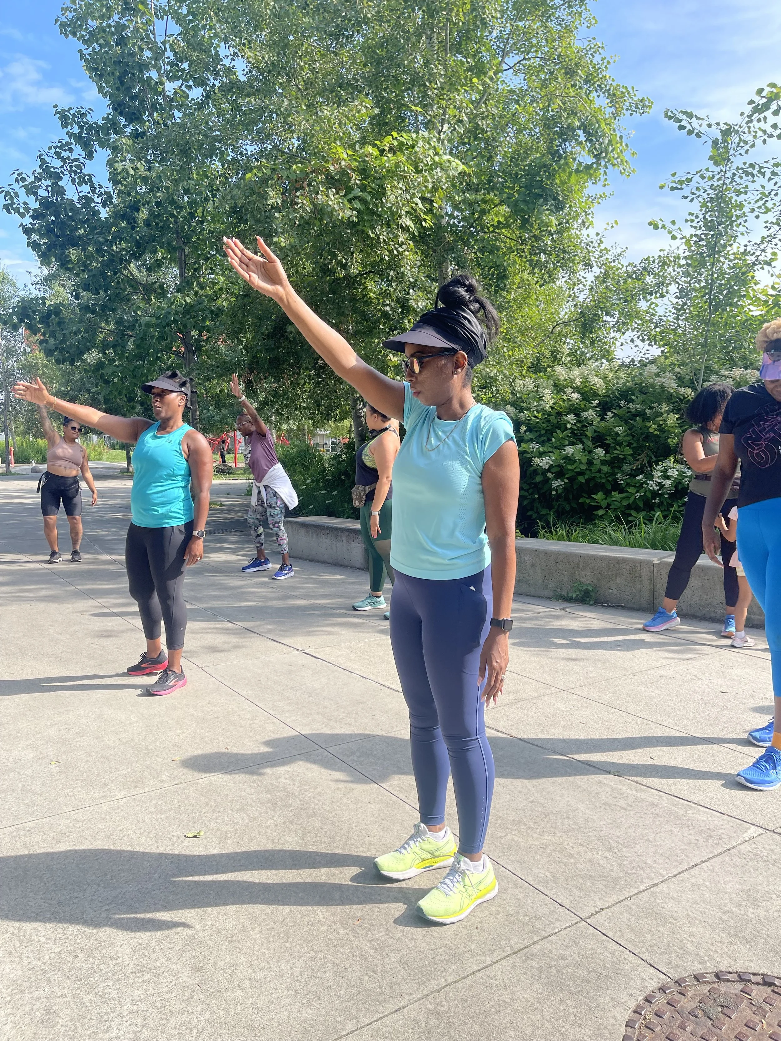 A group of women participating in outdoor exercise or stretching in a park on a sunny day, with trees and greenery in the background.