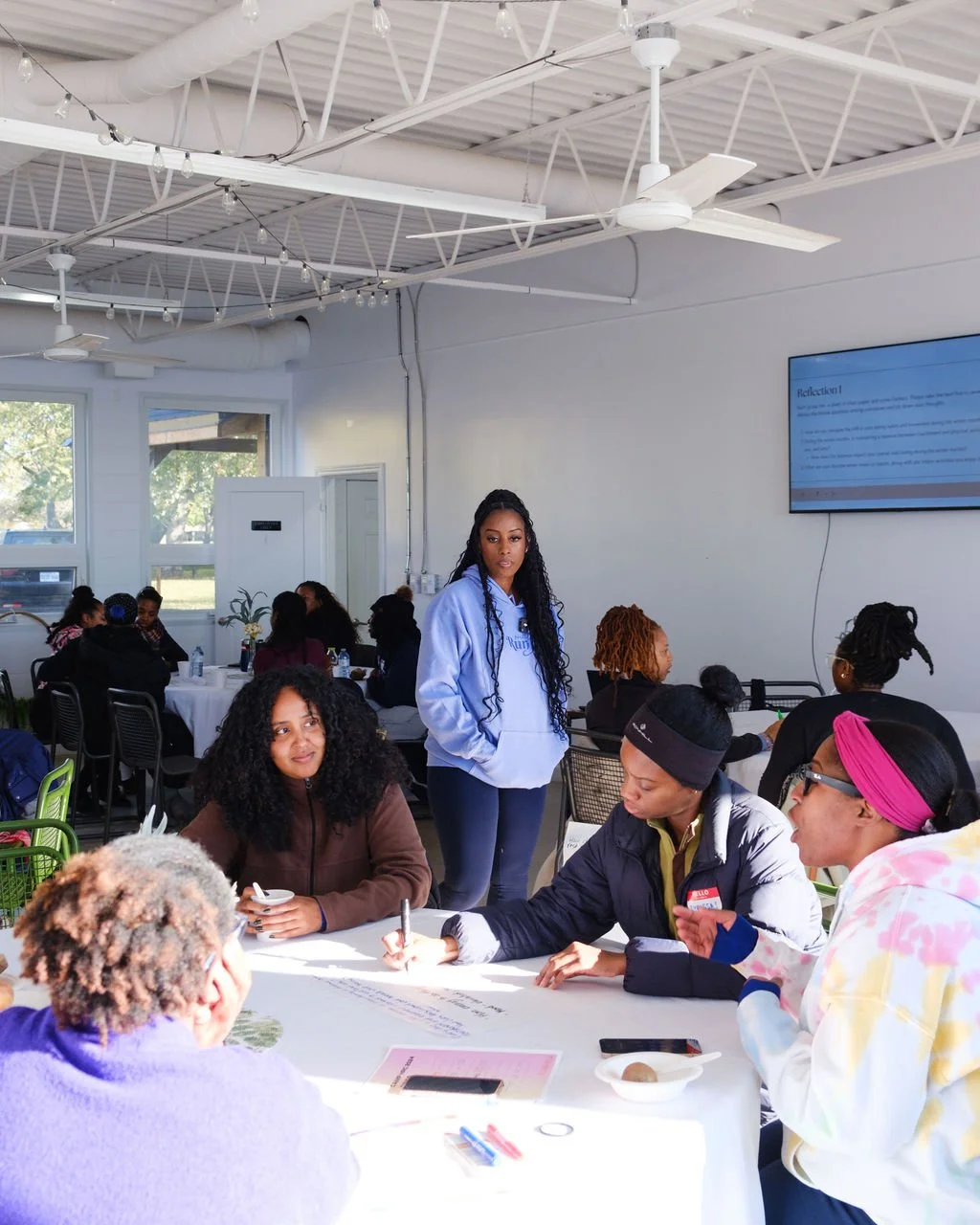 A group of diverse women sitting around a table in a bright, modern room, participating in a discussion or workshop, with one woman standing near a wall-mounted screen.