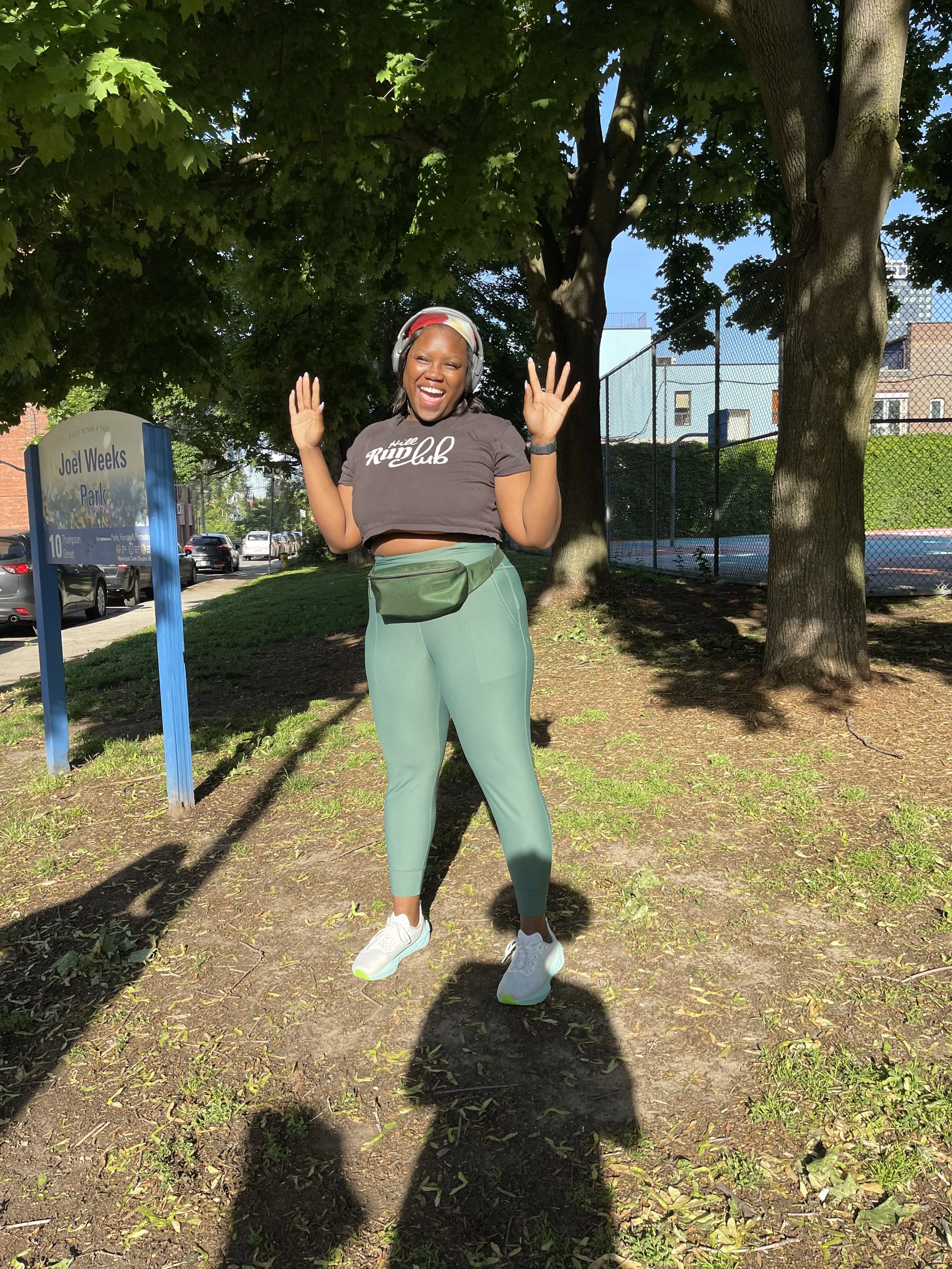 A woman with long dreadlocks smiling and jumping in a park, wearing a brown t-shirt, light green leggings, white sneakers, a green waist bag, and headphones, with trees and a fence in the background.