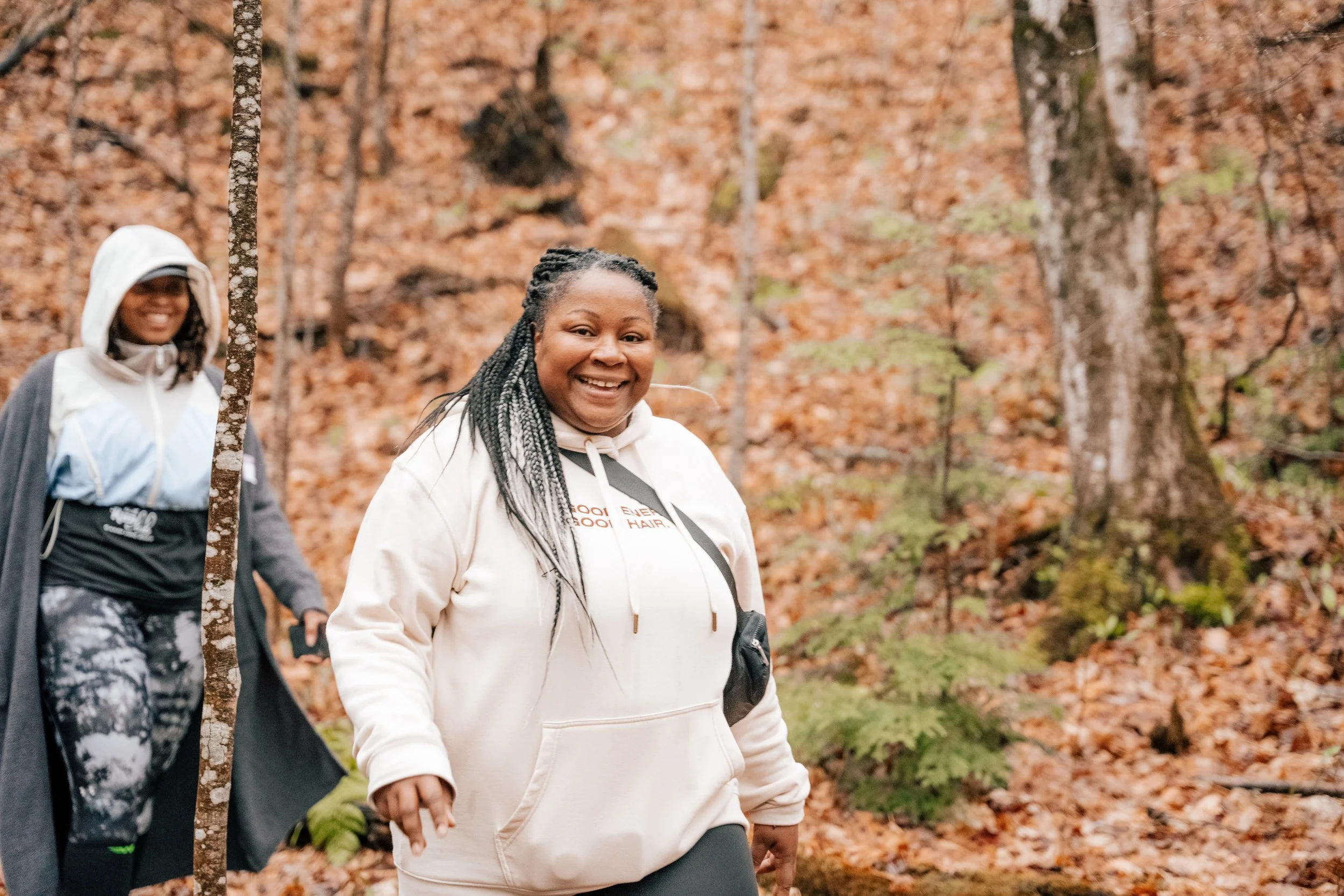 Two women hiking through a forest with orange and green leaves on trees and ground, smiling at the camera.
