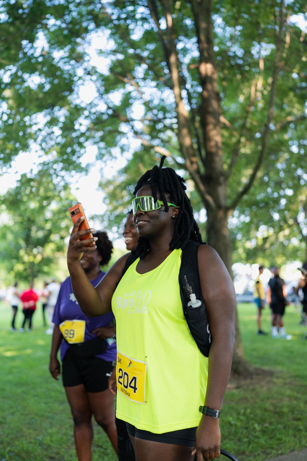 A woman with dreadlocks wearing a neon yellow running tank top, sunglasses, and a black backpack, holding a smartphone and smiling at an outdoor event with trees and other runners in the background.