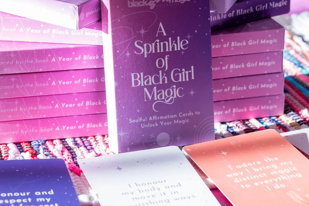 Display of purple boxes titled "A Sprinkle of Black Girl Magic" with soul affirmation cards in pink, purple, and white, arranged on a multicolored woven surface.
