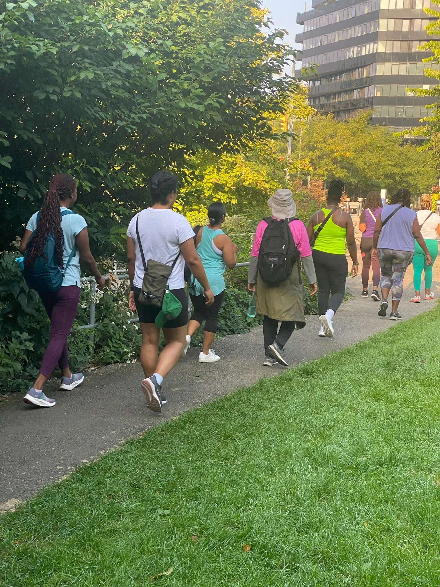 Group of women walking together in a park on a sunny day, with trees and modern buildings in the background.