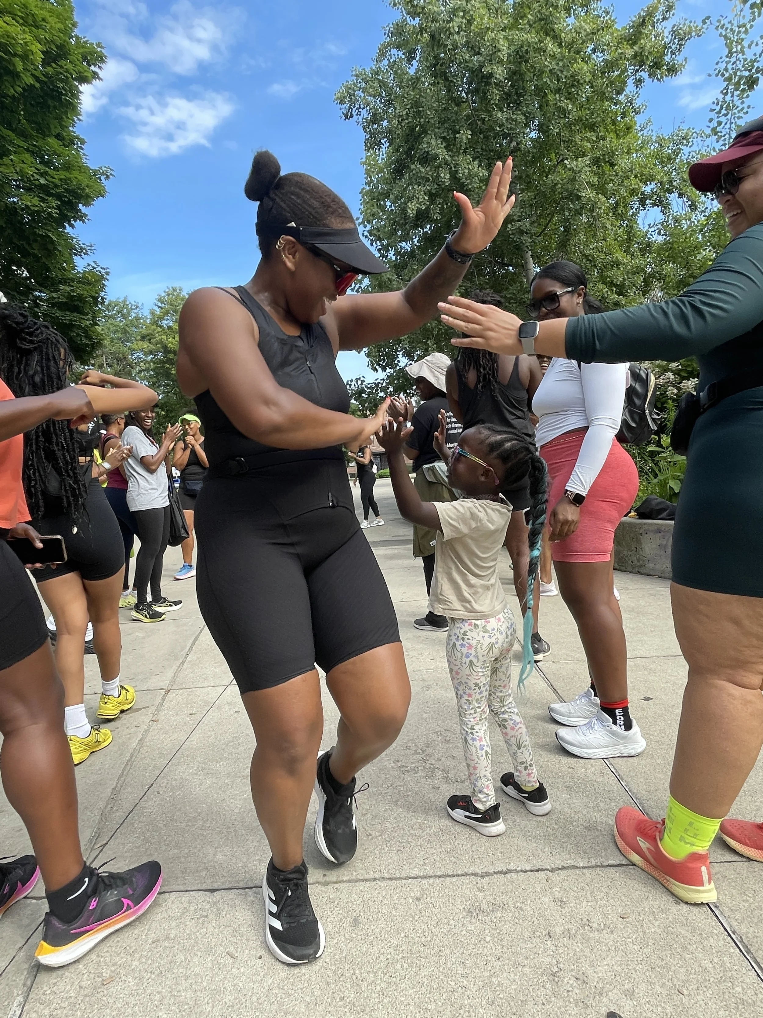 A group of people dancing outdoors on a sunny day, with a woman in black shorts and a visor leading a dance with a young girl, surrounded by smiling onlookers.