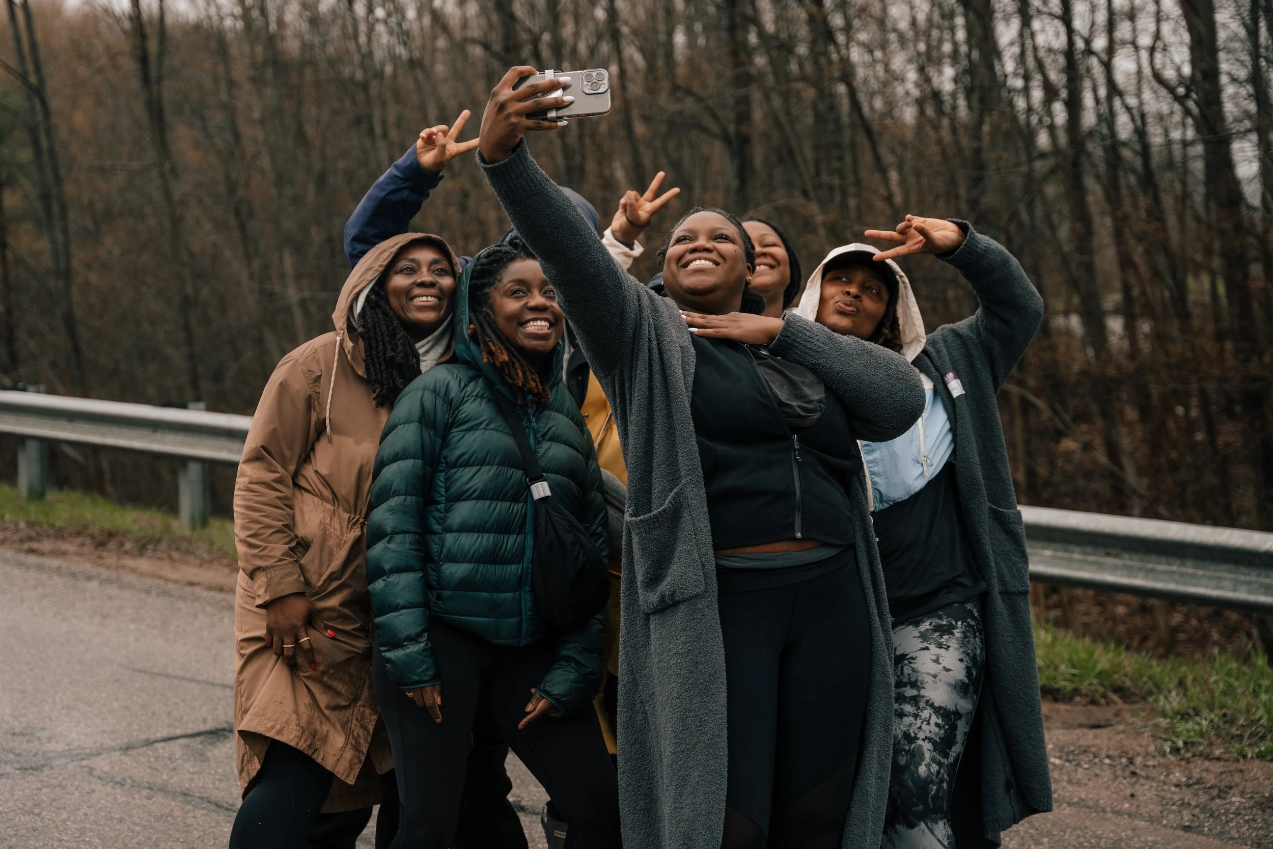 Group of women taking a selfie outdoors on a cloudy day in a wooded area, wearing jackets and making peace signs.