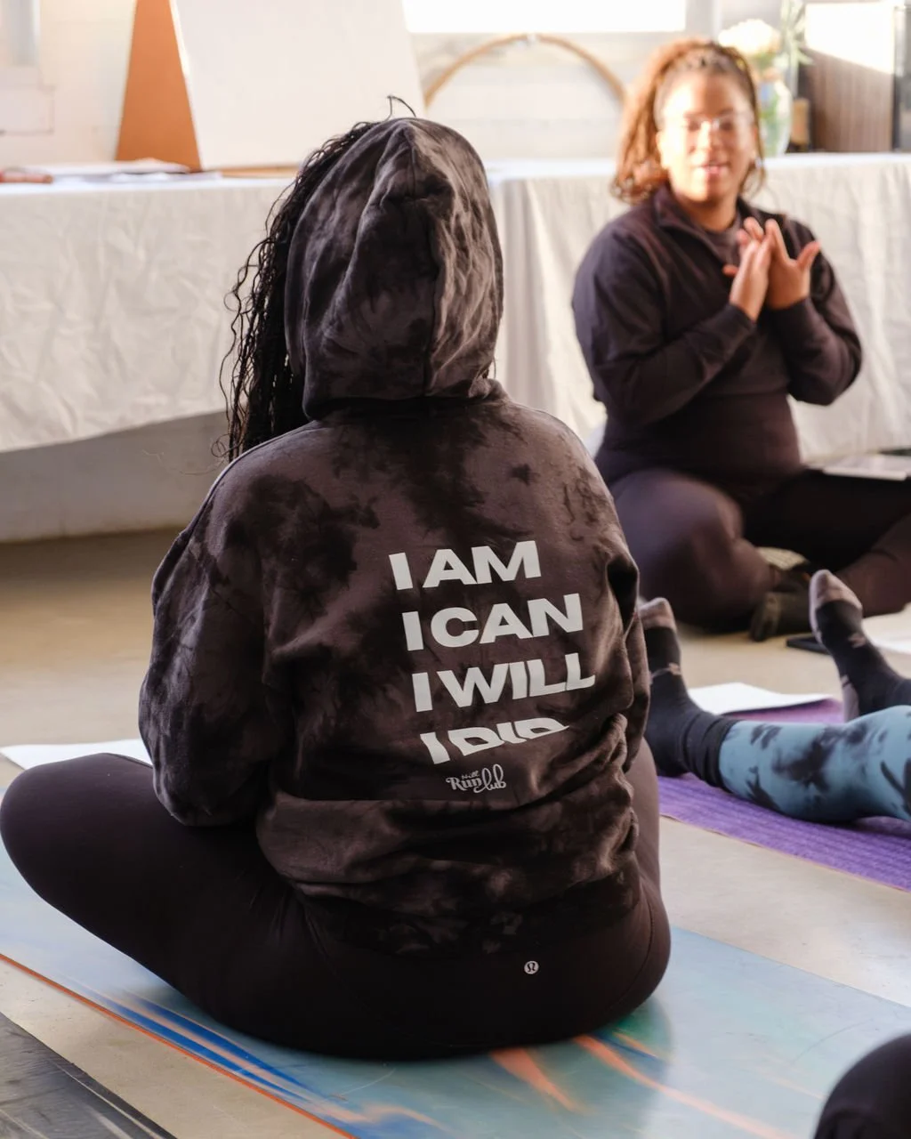 A woman in a black hoodie with the words "I AM I CAN I WILL I DO" sitting cross-legged on a yoga mat, facing another woman in a black jacket, sitting on the floor with hands on her chest, in a room with a white table and potted plant in the backgroun