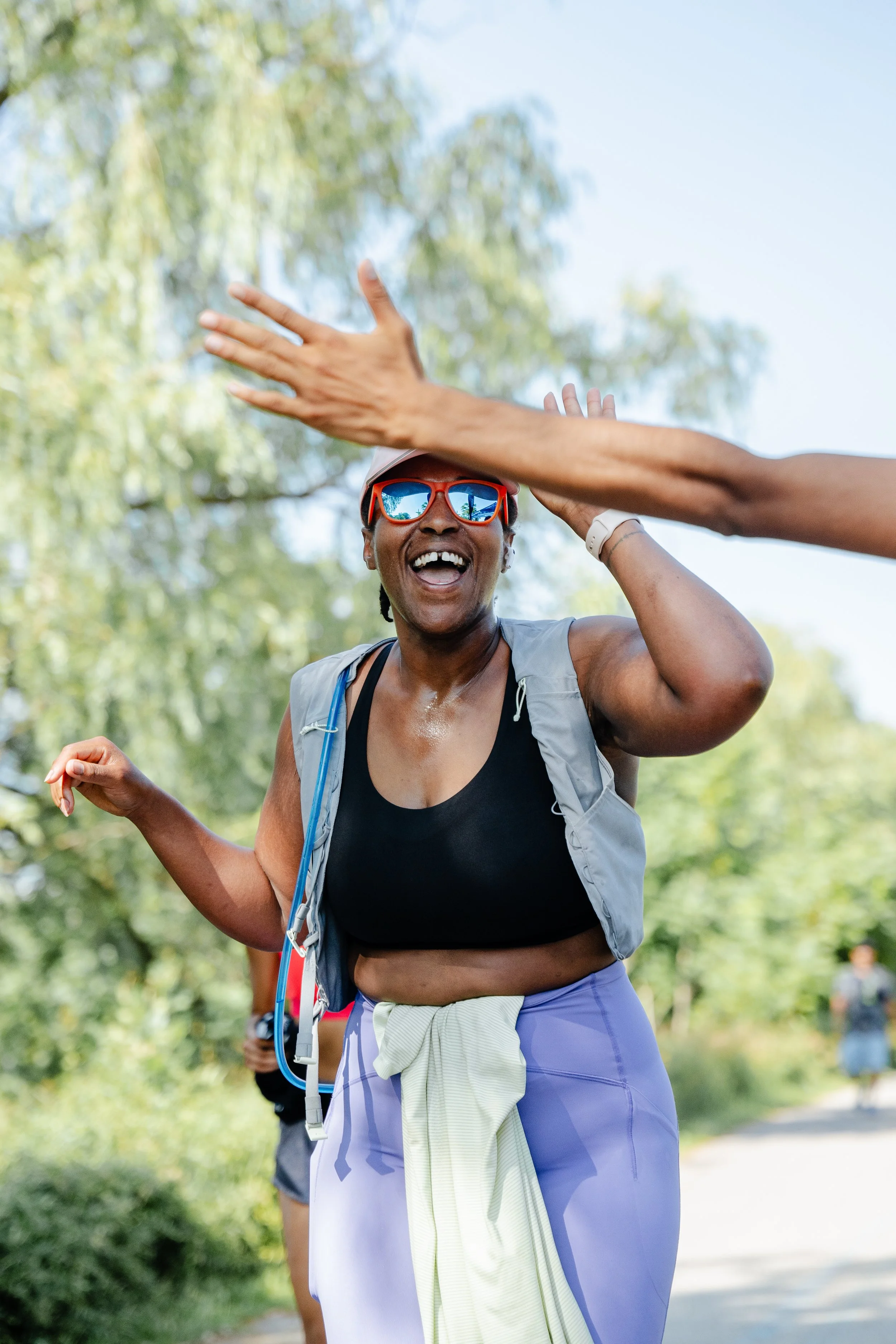 Woman smiling and celebrating outdoors, wearing sunglasses, sports bra, and workout gear, with another person's hand reaching towards her.