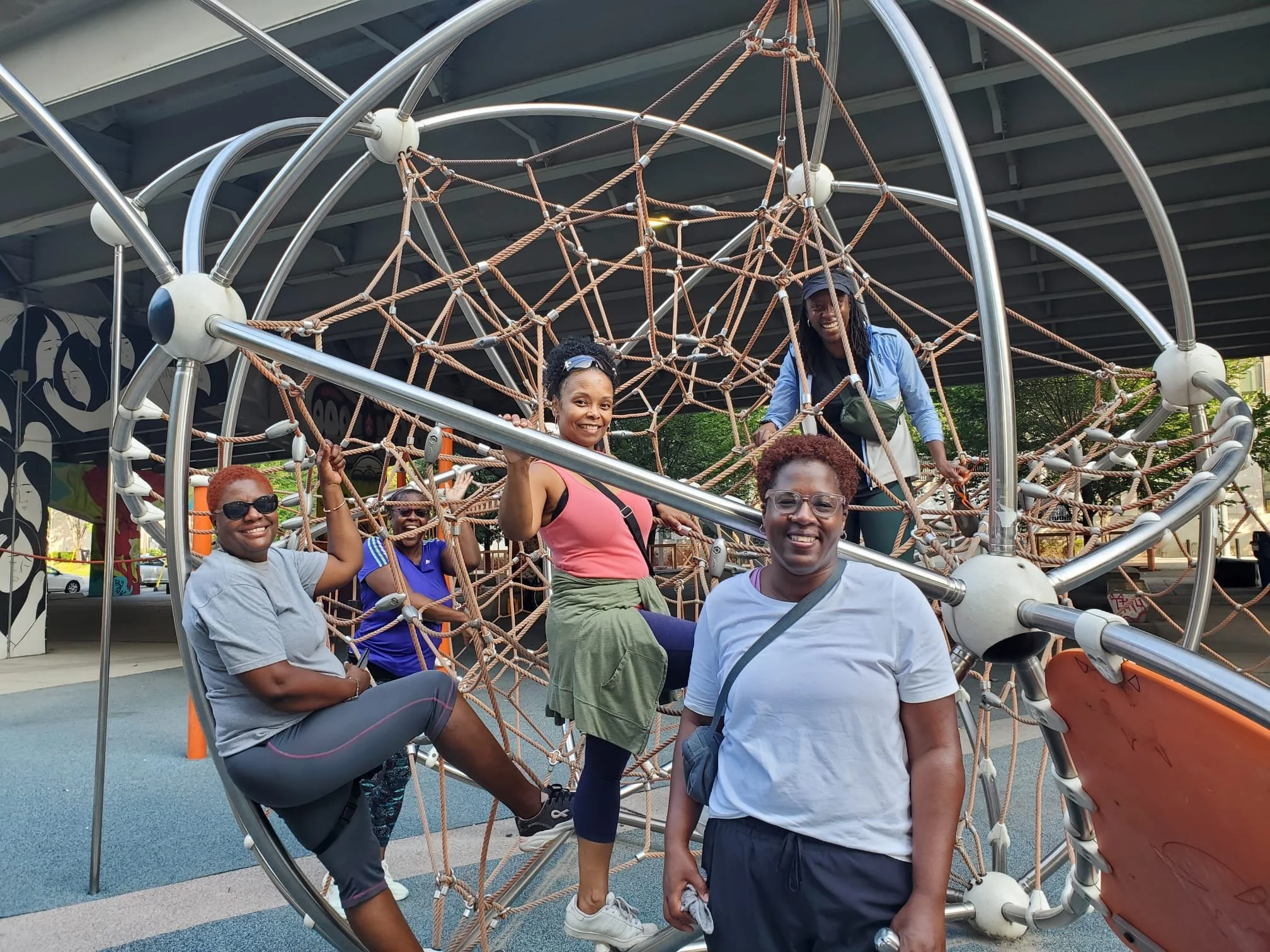 Group of five women at a playground, climbing on a large, spherical rope climbing structure under a bridge.