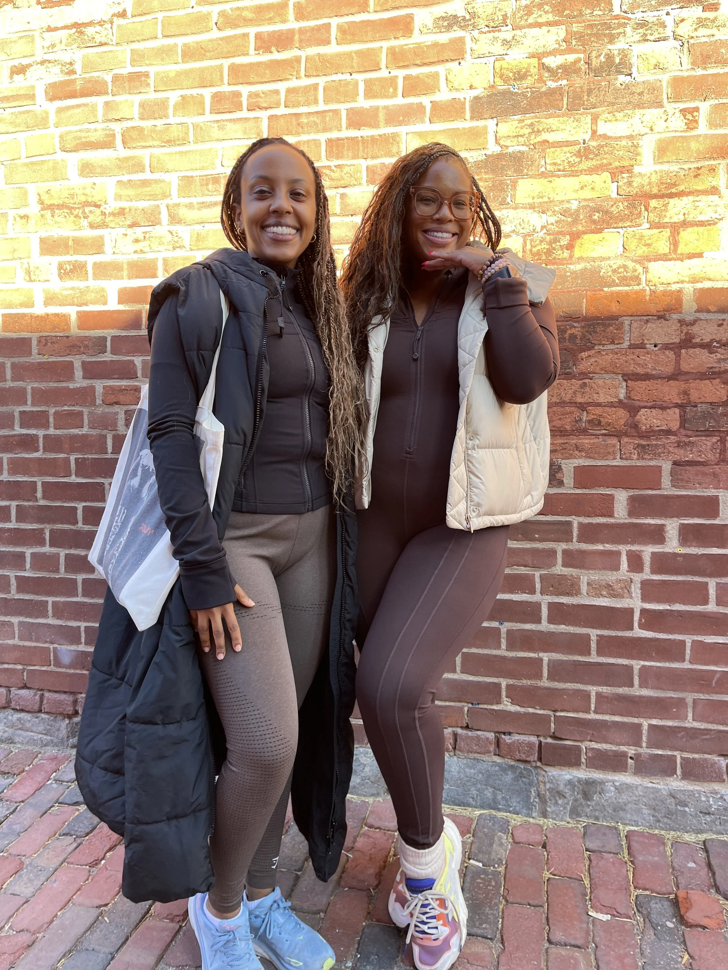Two women standing against a brick wall, smiling at the camera, dressed in athletic wear and jackets.