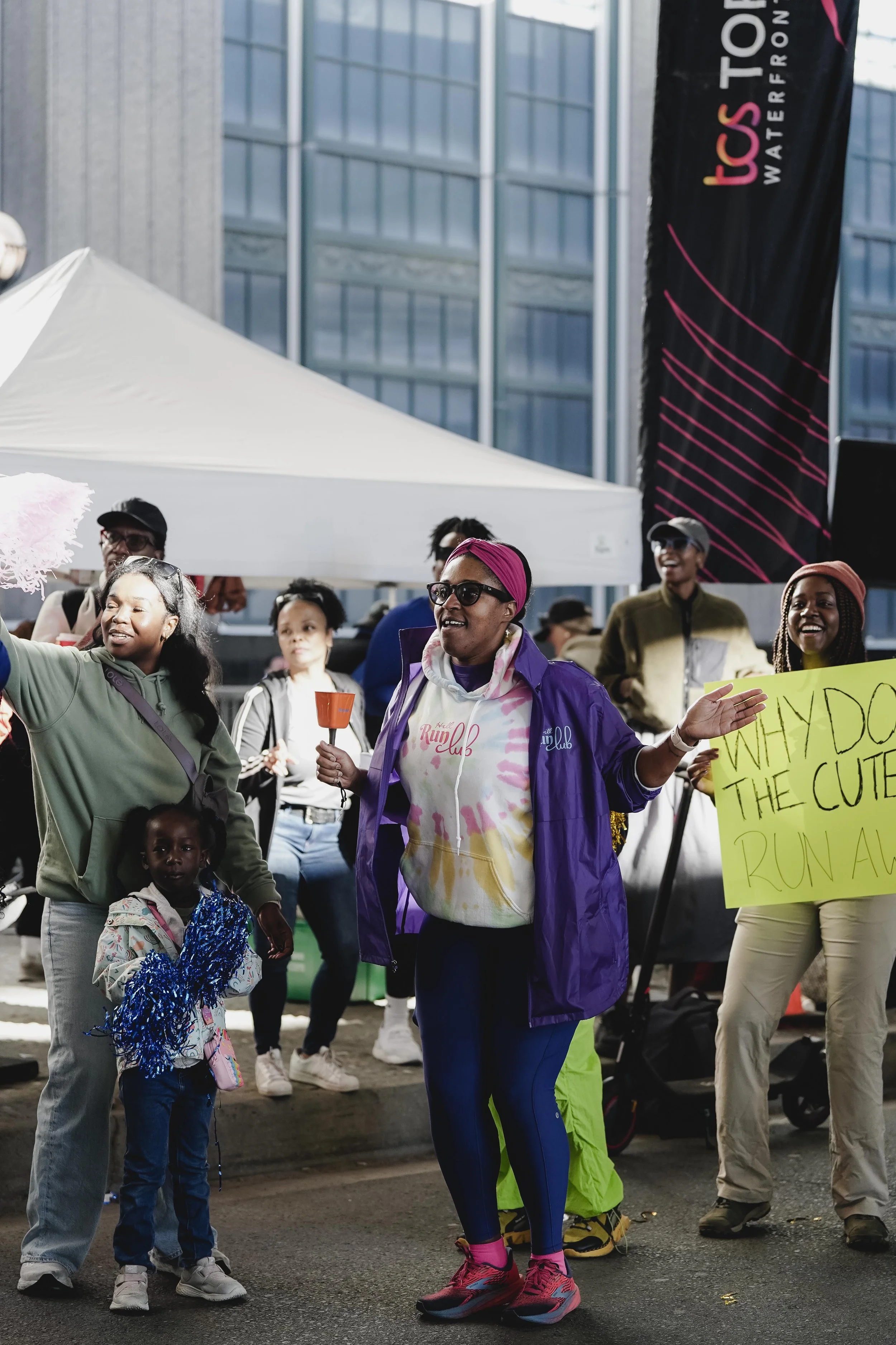 Group of diverse women and children at an outdoor event, with one woman holding a sign that says 'WHY DO THE CUTES RUN AWAY,' and another woman holding a small orange bell, all smiling and engaging in festivities.
