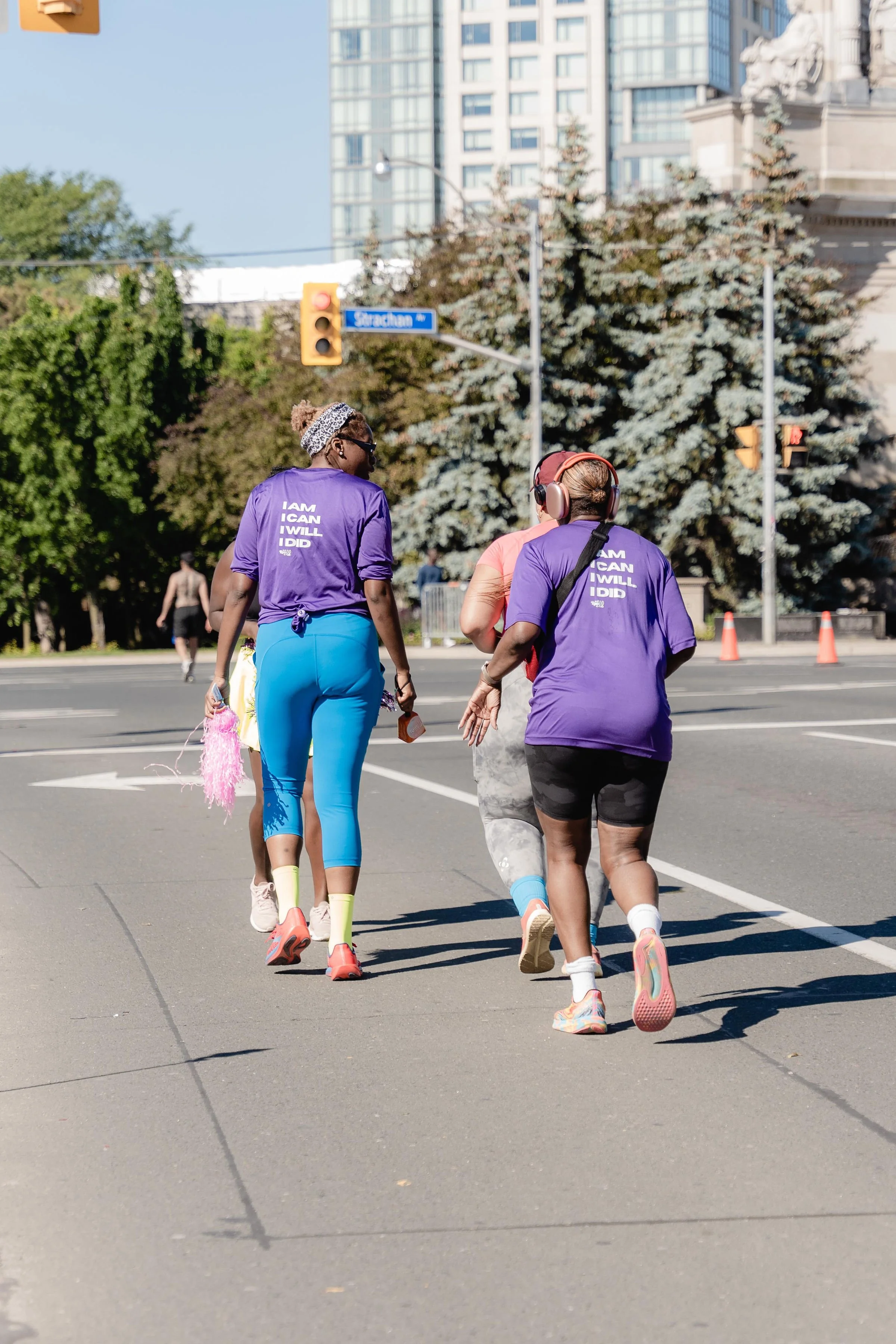 People jogging and walking across a city street, with trees and tall buildings in the background.